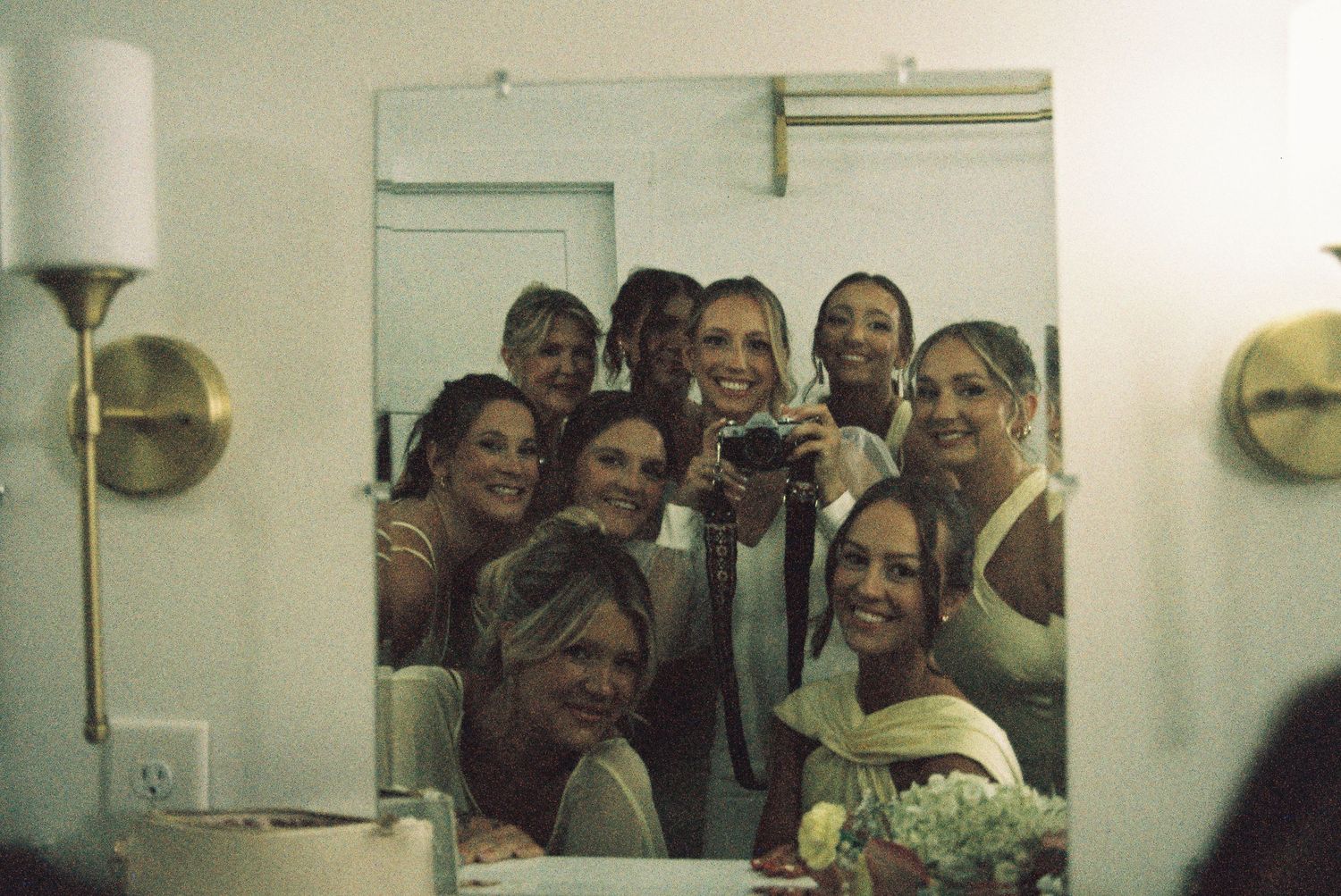 Group of bridesmaids and bride smiling together in mirror reflection during wedding day getting ready.