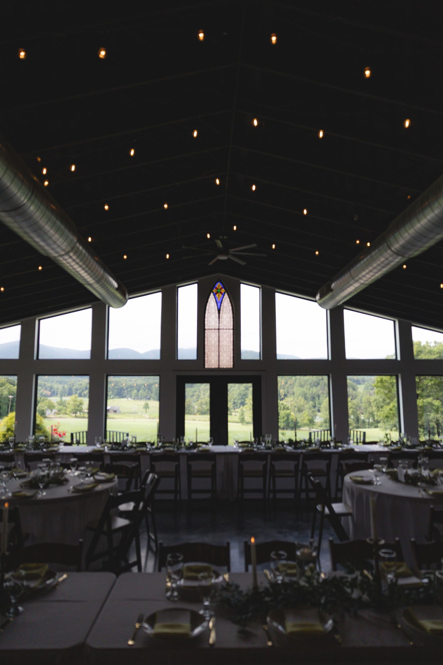 Elegant wedding reception hall with string lights, stained glass window, round tables, and scenic mountain views.
