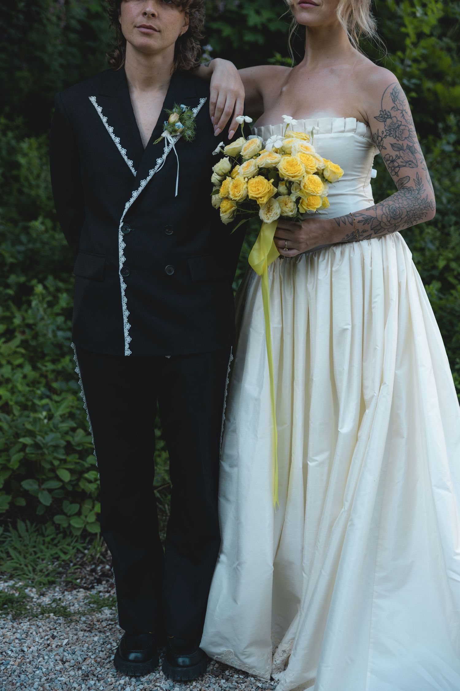 Couple in wedding attire, groom in black suit, tattooed bride in white strapless gown holding yellow rose bouquet.