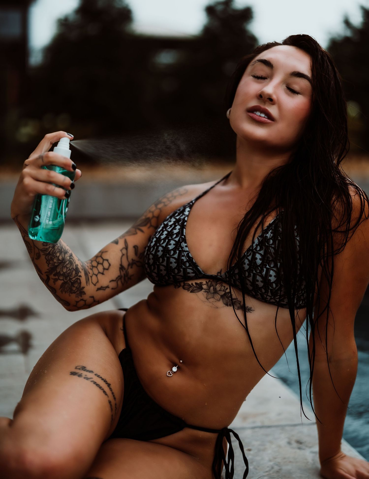 Tattooed woman in black bikini spraying mist, with wet hair and belly piercing, posing outdoors.