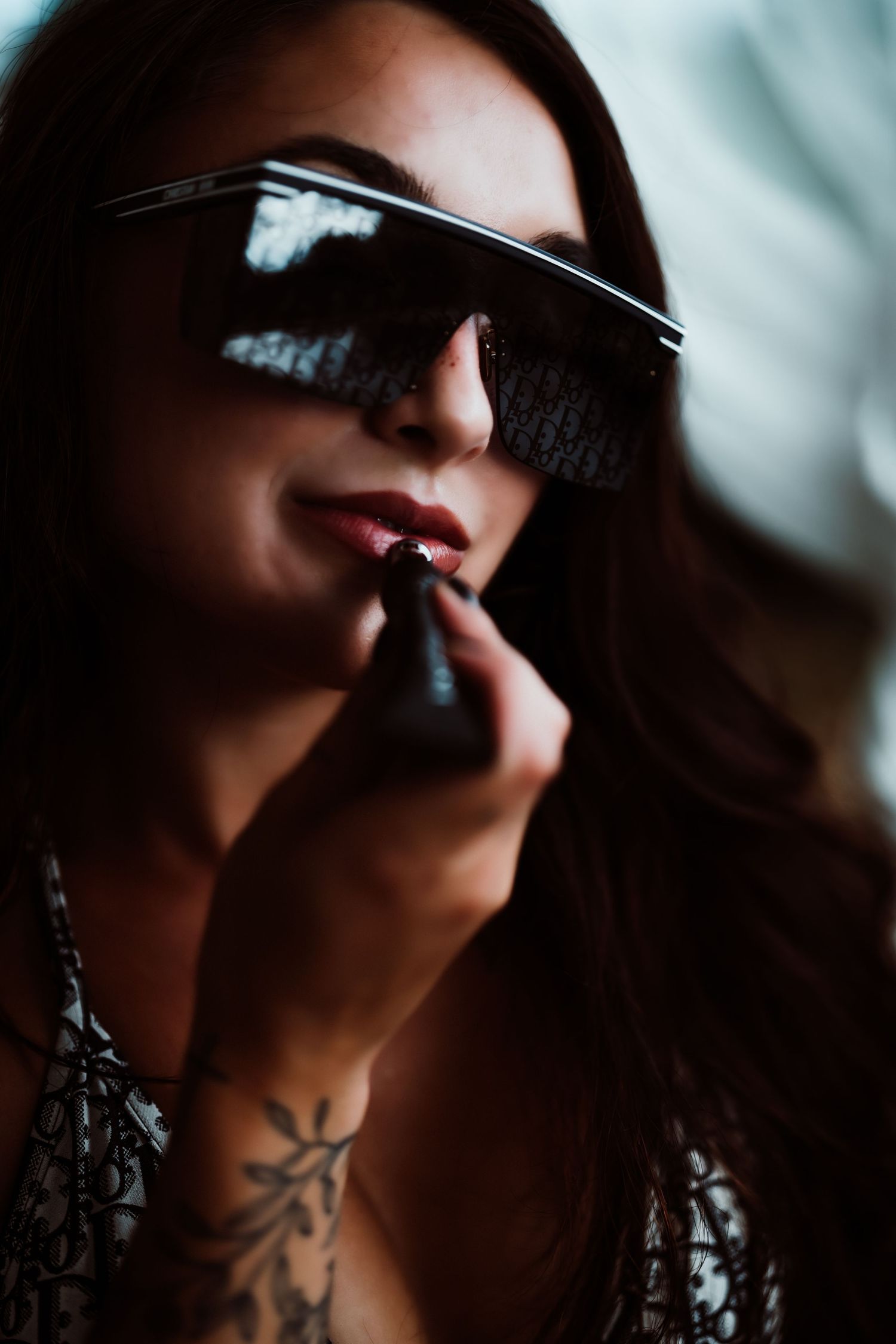 Woman with tattoo applying red lipstick wearing stylish dark square sunglasses with long brunette hair.