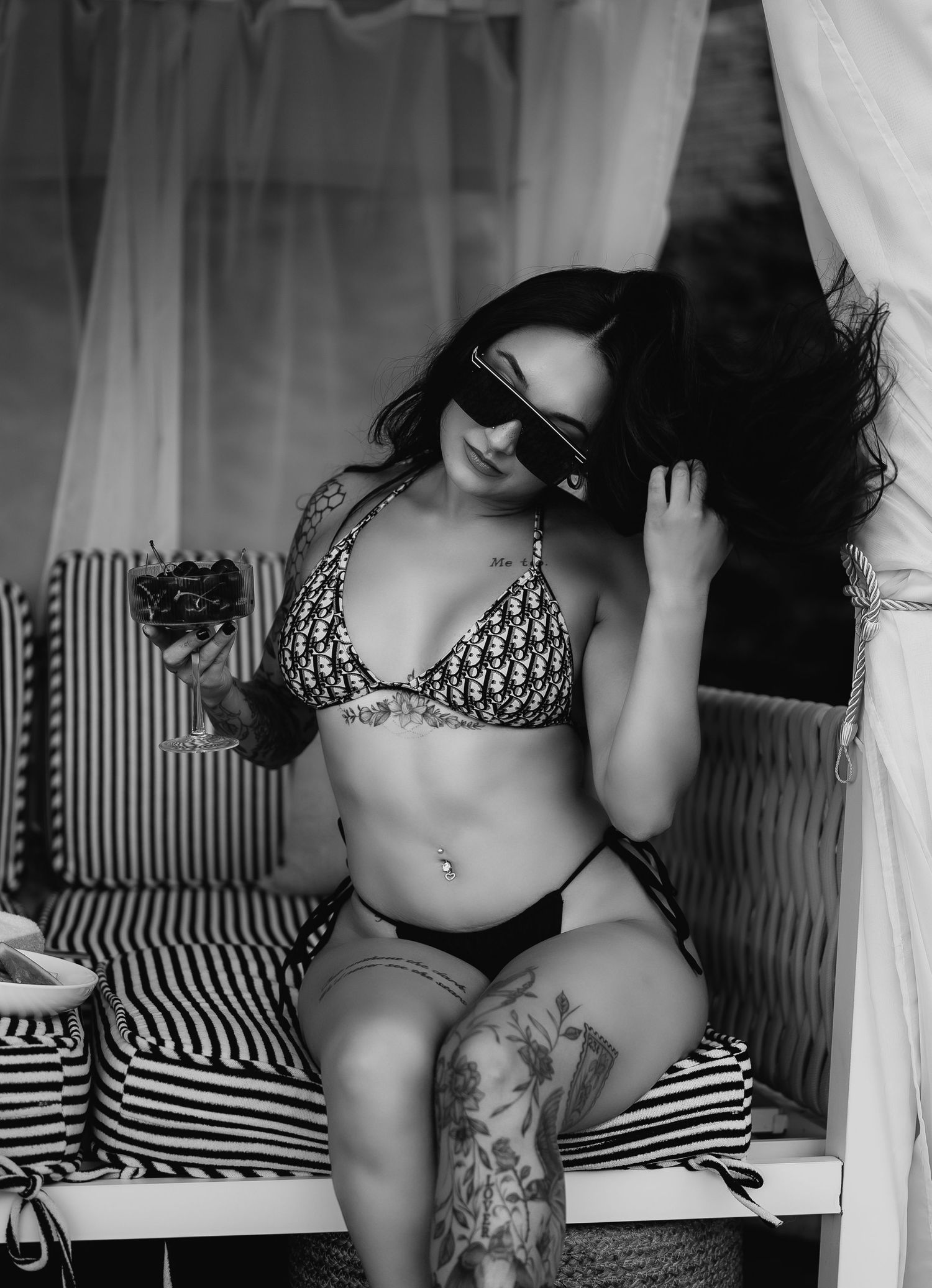 Black and white photo of tattooed woman in bikini and sunglasses holding a drink in a cabana.