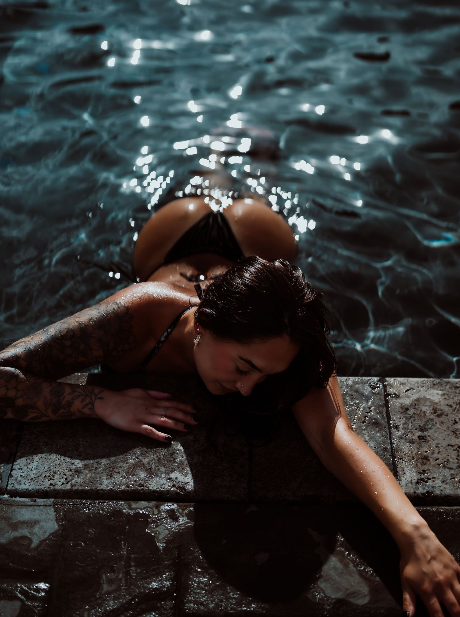 Tattooed woman in black bikini relaxing at pool edge with sparkling water in background.