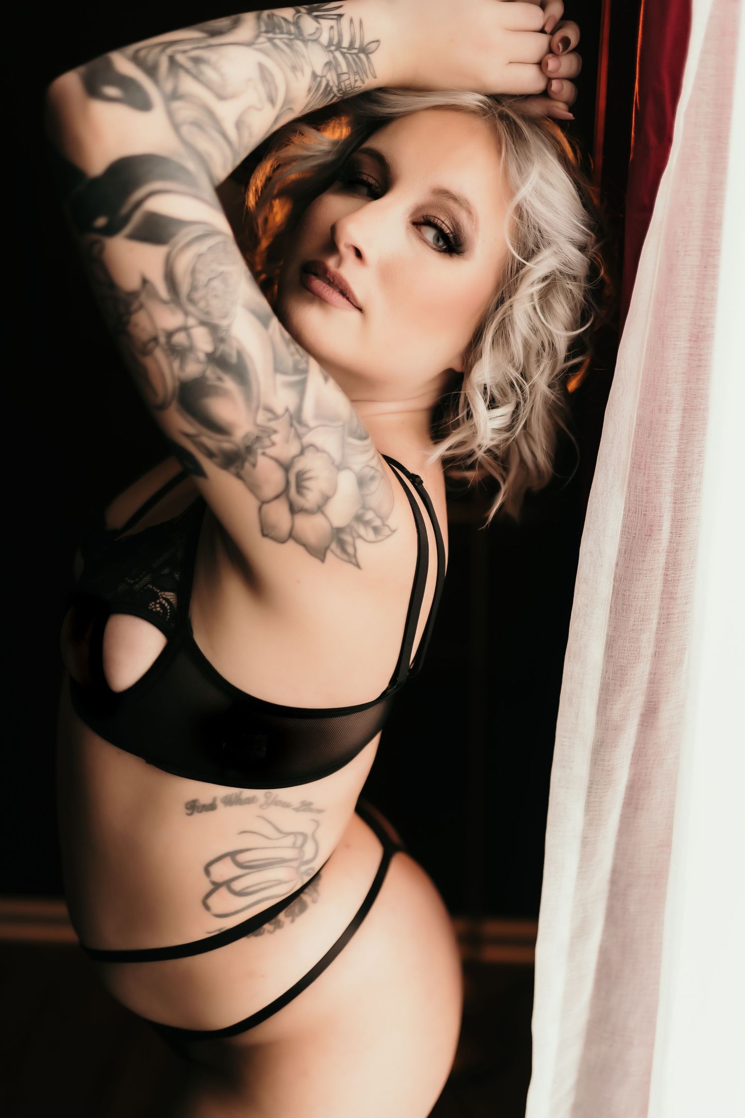 Tattooed blonde woman posing in black lingerie near sheer curtain in dramatic boudoir photography.