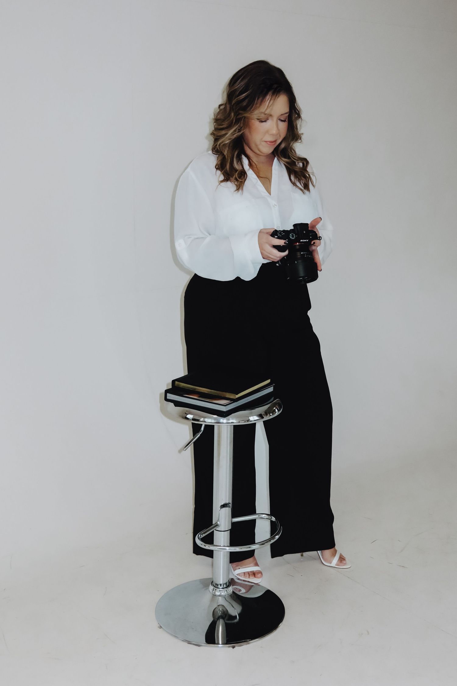 Woman in white blouse and black pants holds camera near chrome barstool with books in a white studio.