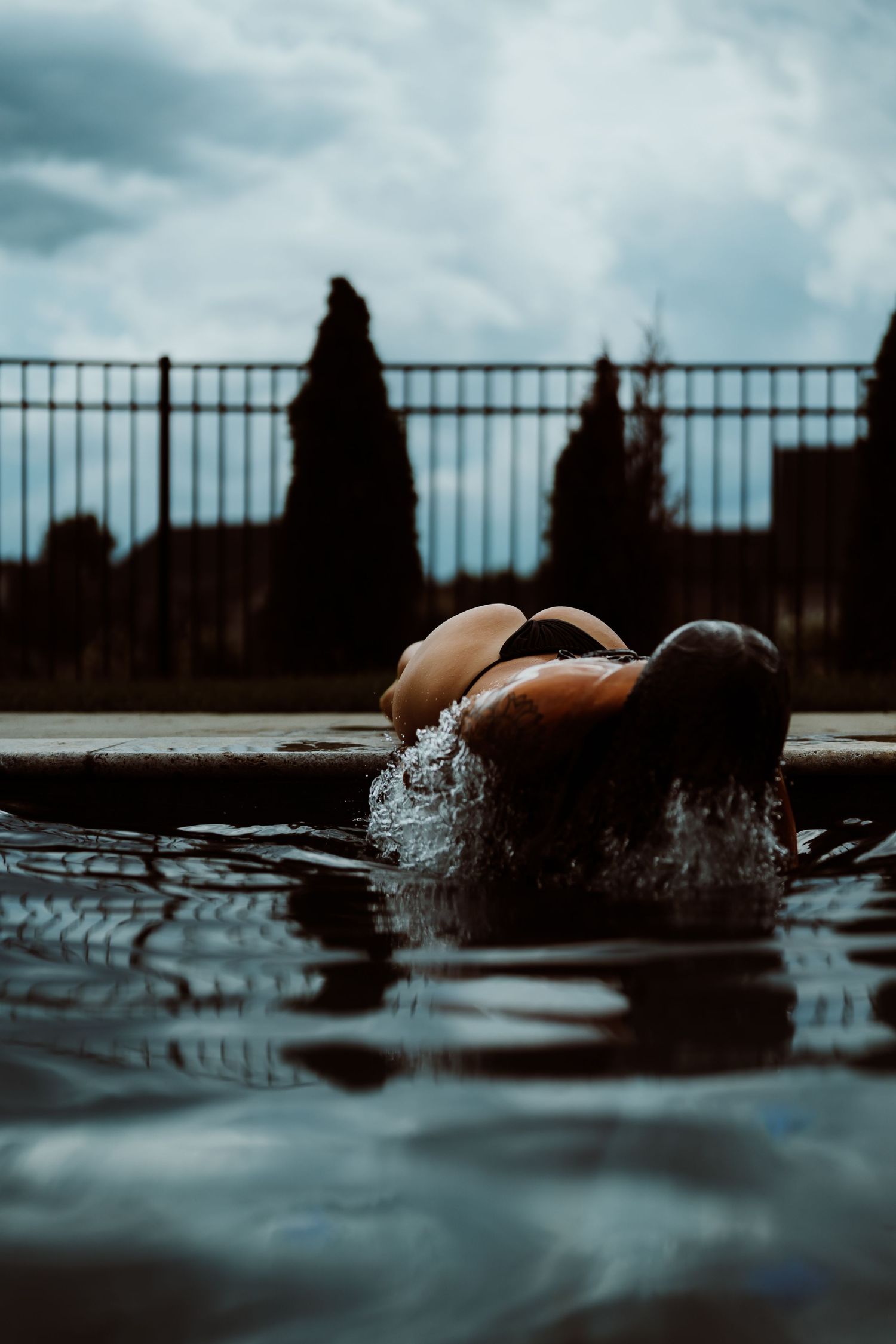 Person splashing in a dark pool with overcast sky, metal fence, and cypress trees in the background.