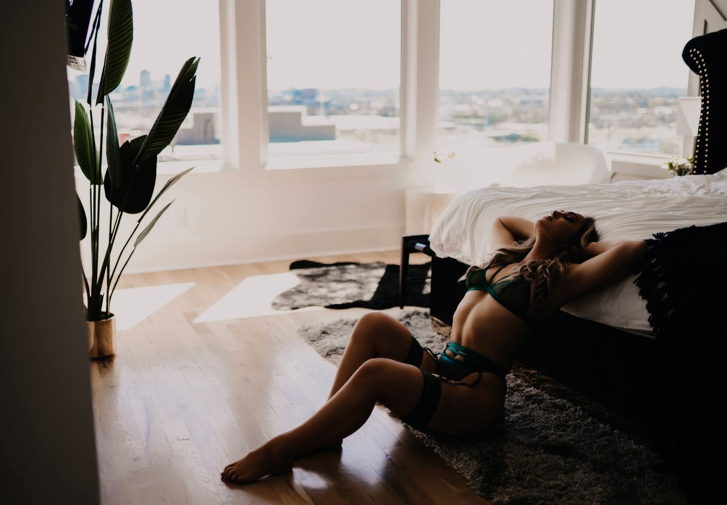 Woman in lingerie relaxing on hardwood floor beside bed near large windows with city view and indoor plants.