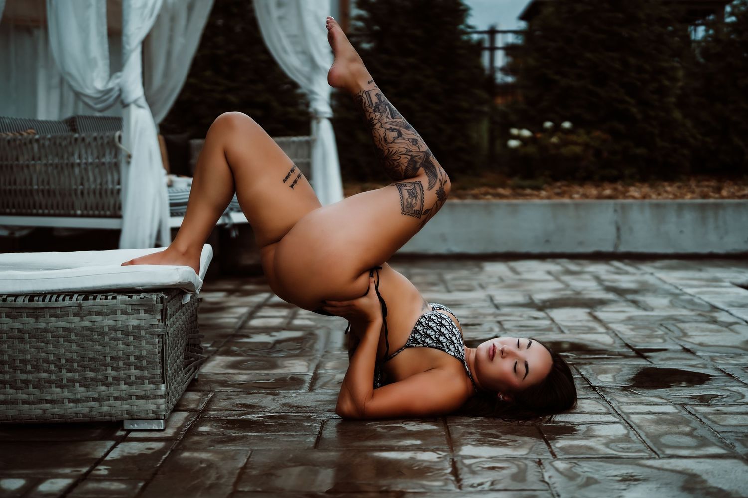 Woman with tattoos posing in bikini on wet tiles outdoors near wicker furniture and white columns.