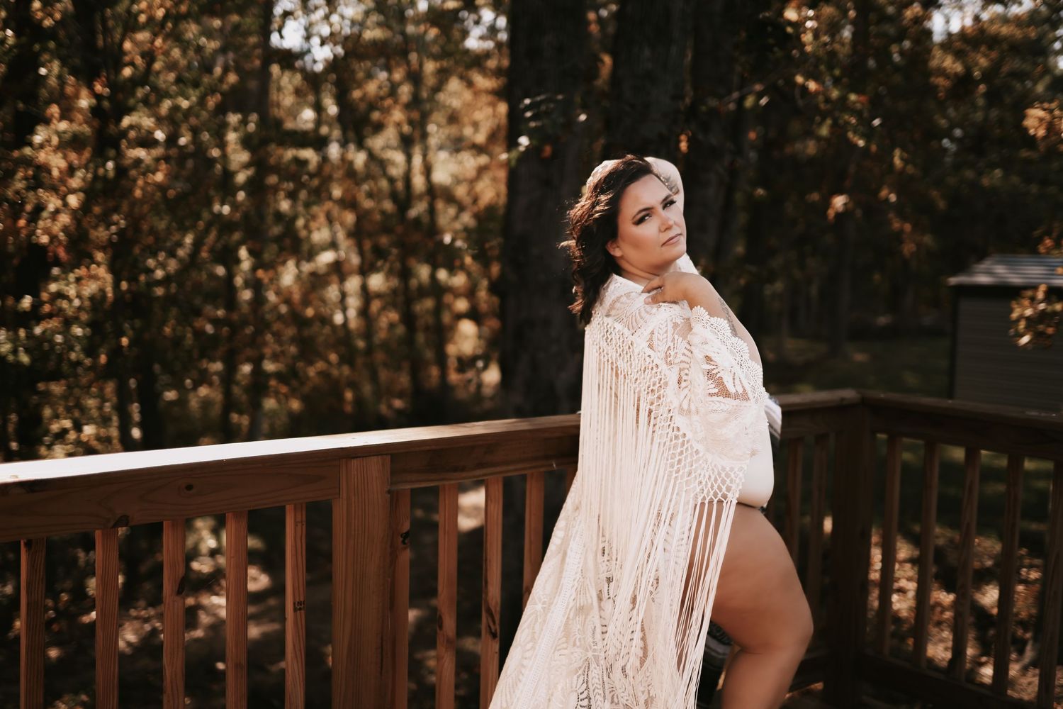 Woman in boho lace wrap poses on wooden deck surrounded by autumn foliage trees.