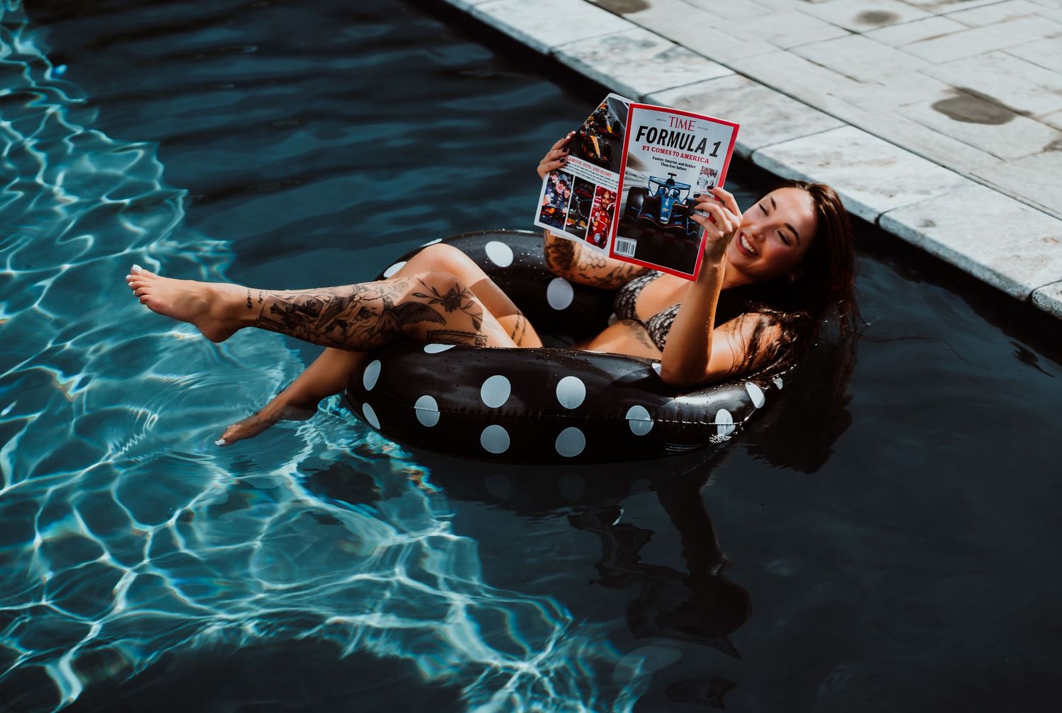 Woman in bikini relaxing on polka dot pool float reading a Formula 1 magazine in a swimming pool.