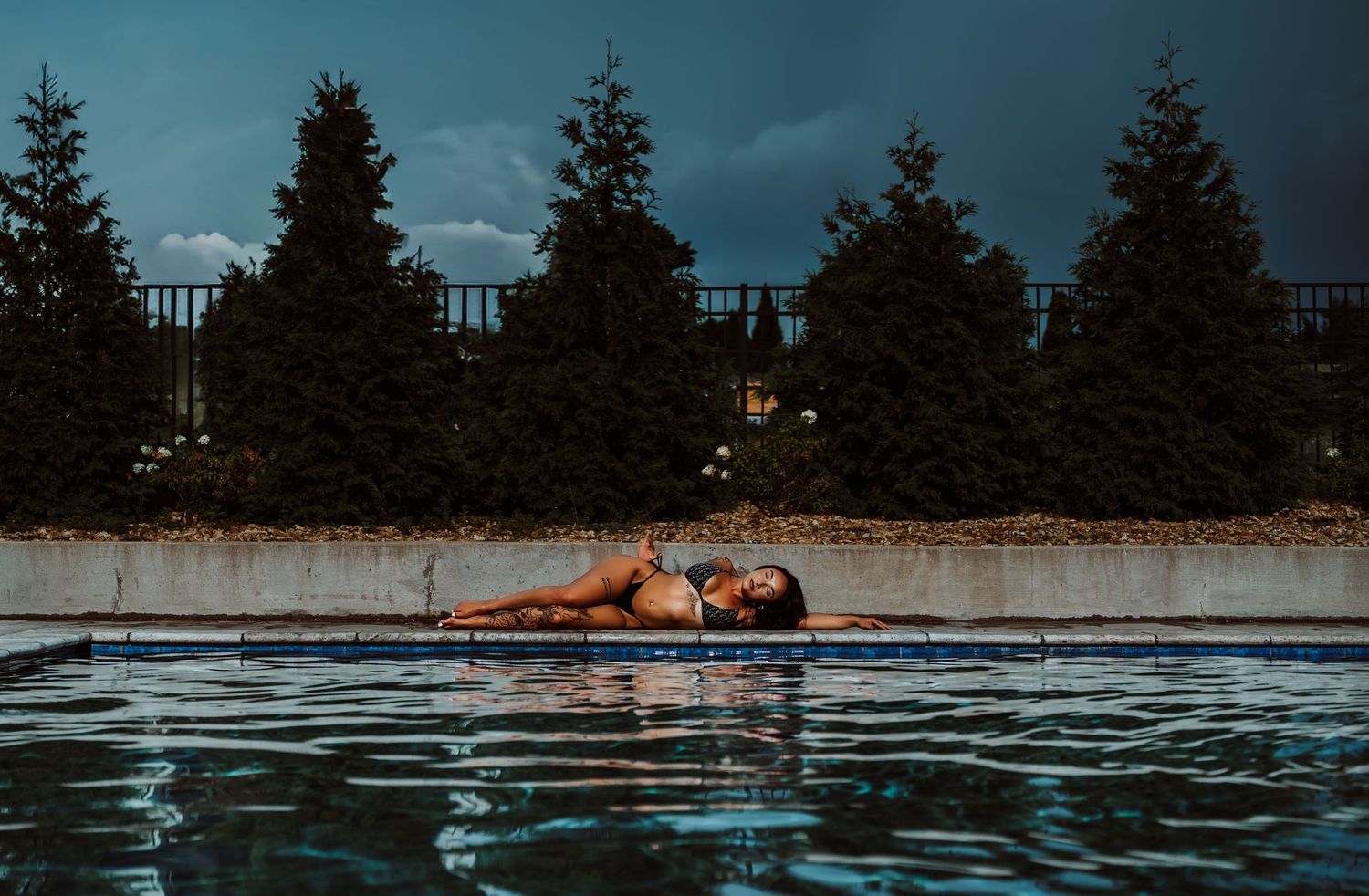 Woman in bikini floating in outdoor pool at dusk with evergreen trees and cloudy sky backdrop.
