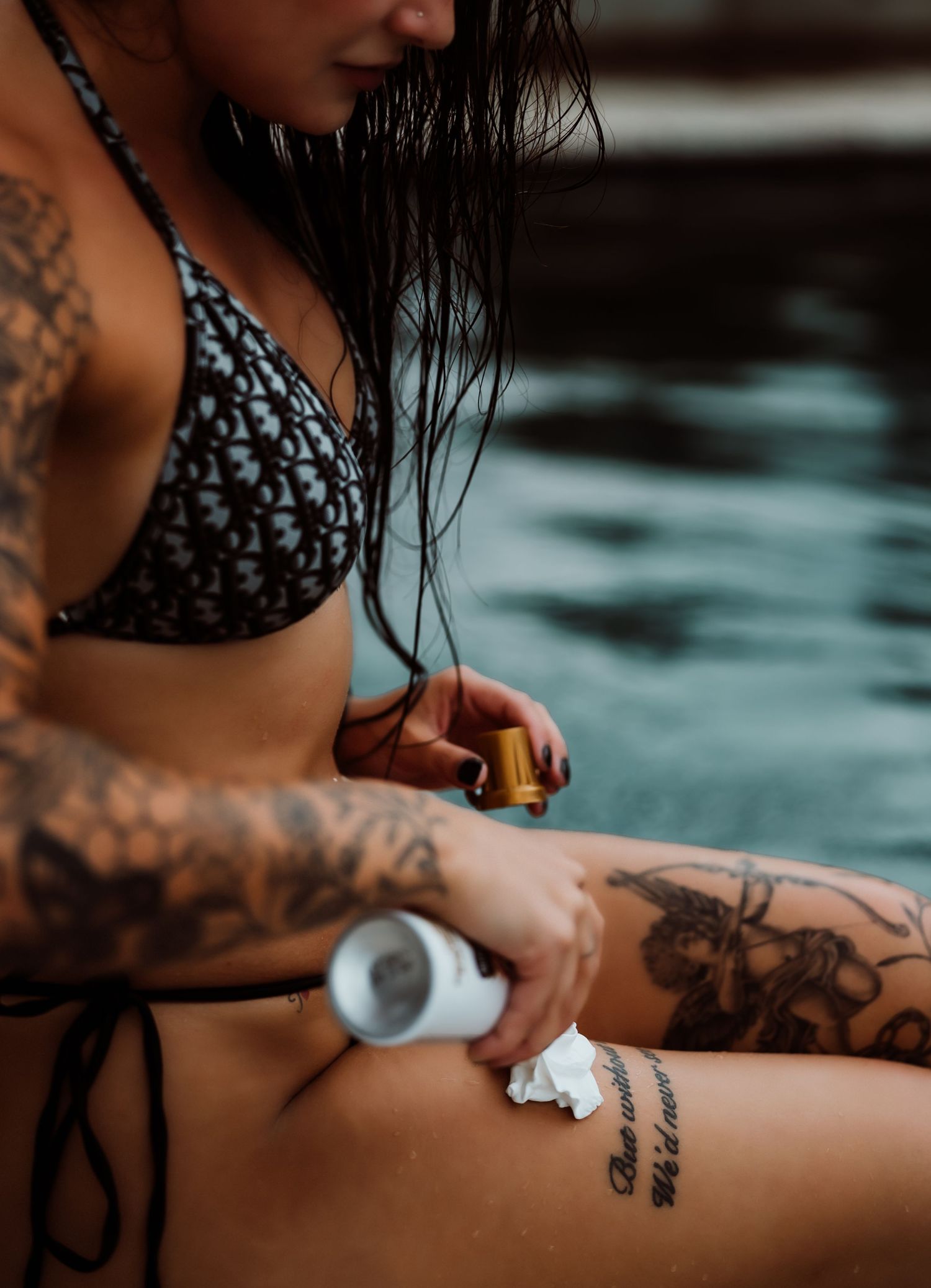 Tattooed woman in bikini applying sunscreen lotion by a pool on a sunny day.