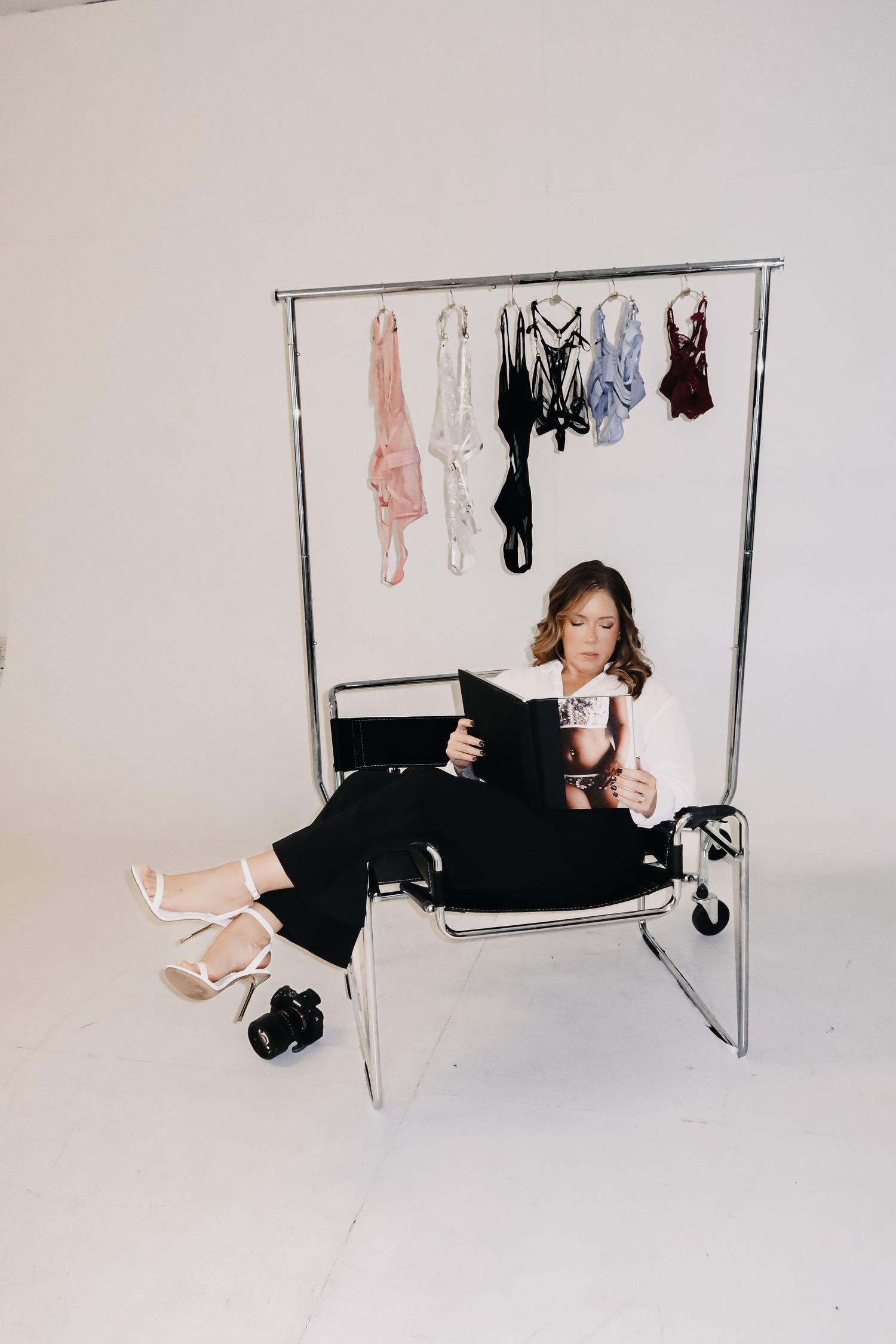 Woman sitting on a chair in a photo studio reviewing a magazine near a clothing rack with lingerie.