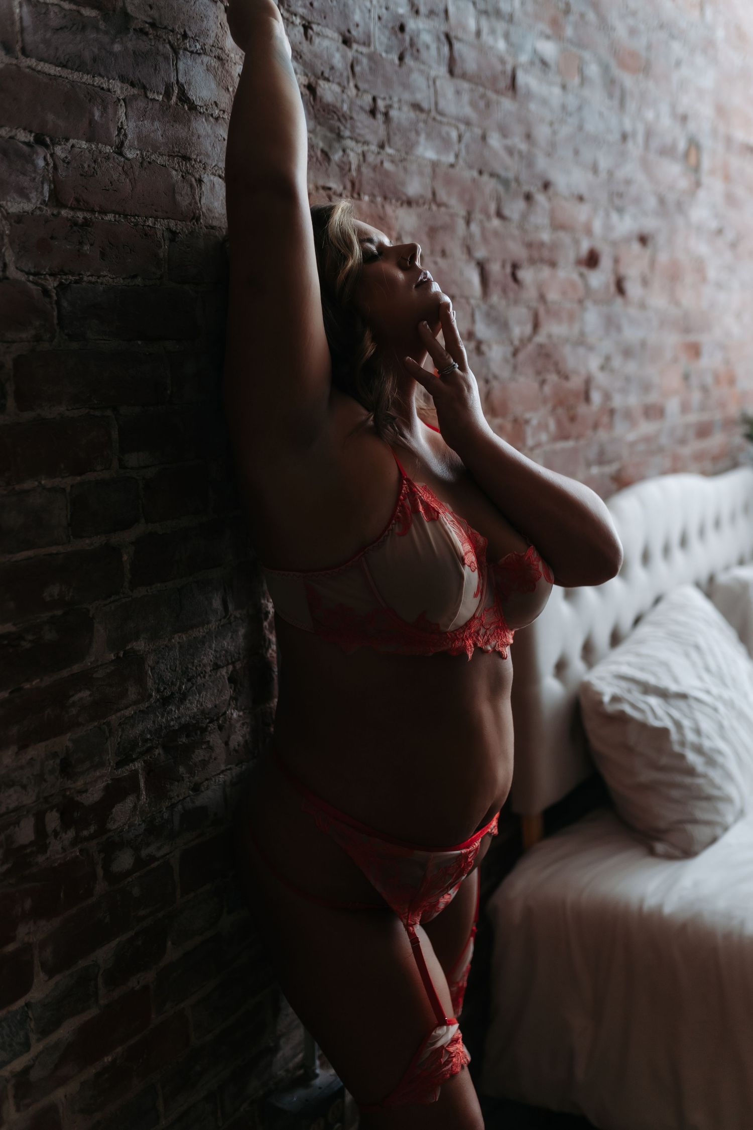 Woman in red lingerie posing against a rustic brick wall near a tufted white sofa in moody lighting.