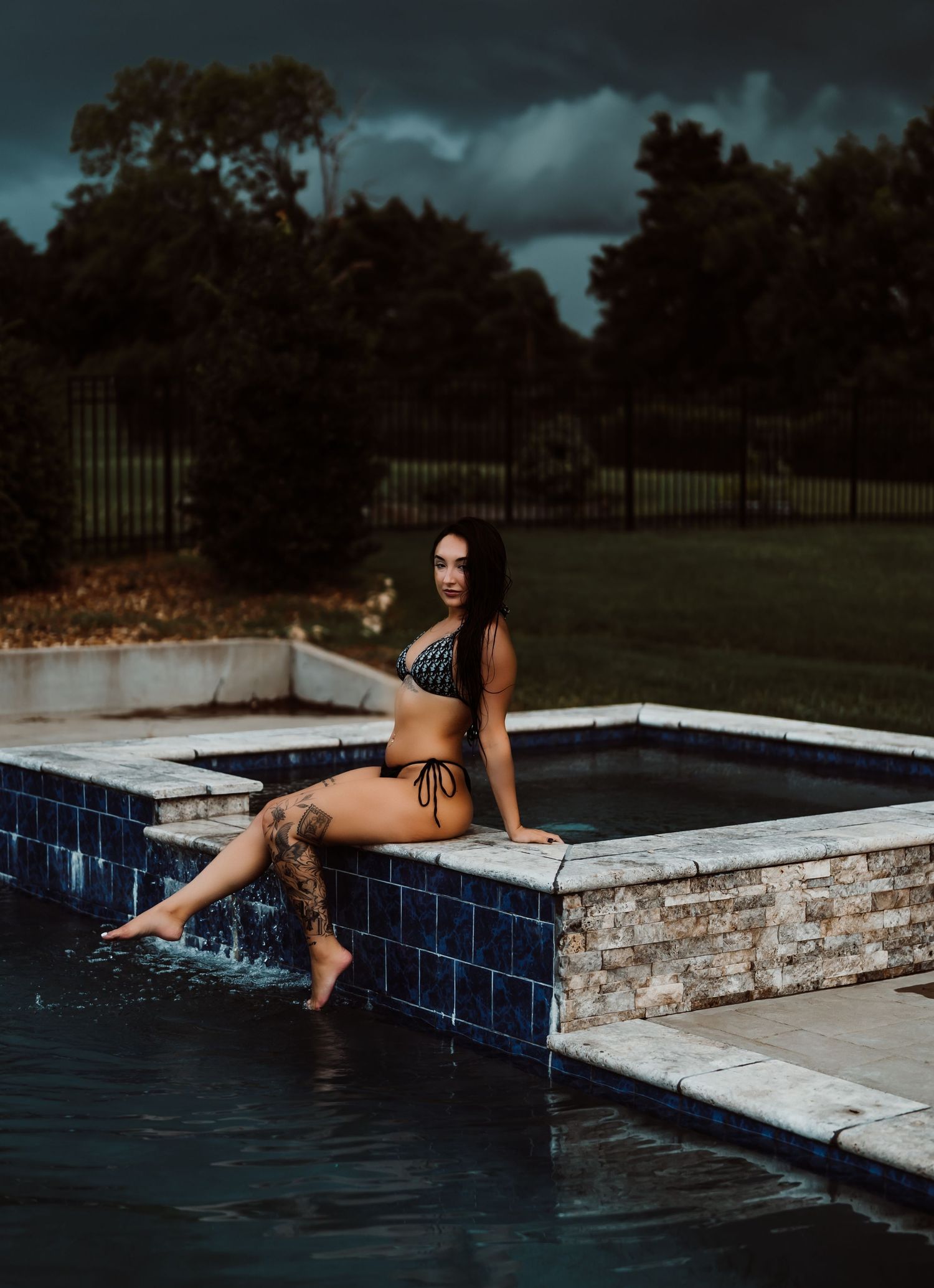 Woman in black bikini sitting poolside with tattoos, stormy sky and lush green trees in background.
