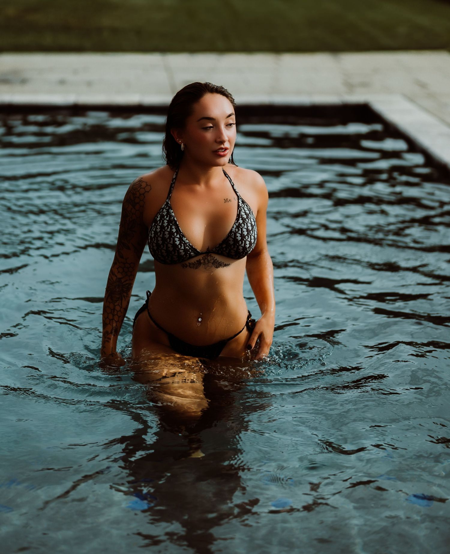 A tattooed woman in a patterned bikini stands waist-deep in a pool, water rippling around her.
