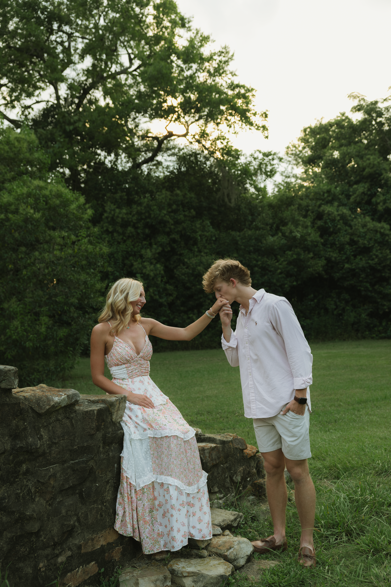 A man kisses a woman's hand outdoors near a stone wall, she wears a floral maxi dress, surrounded by lush greenery.