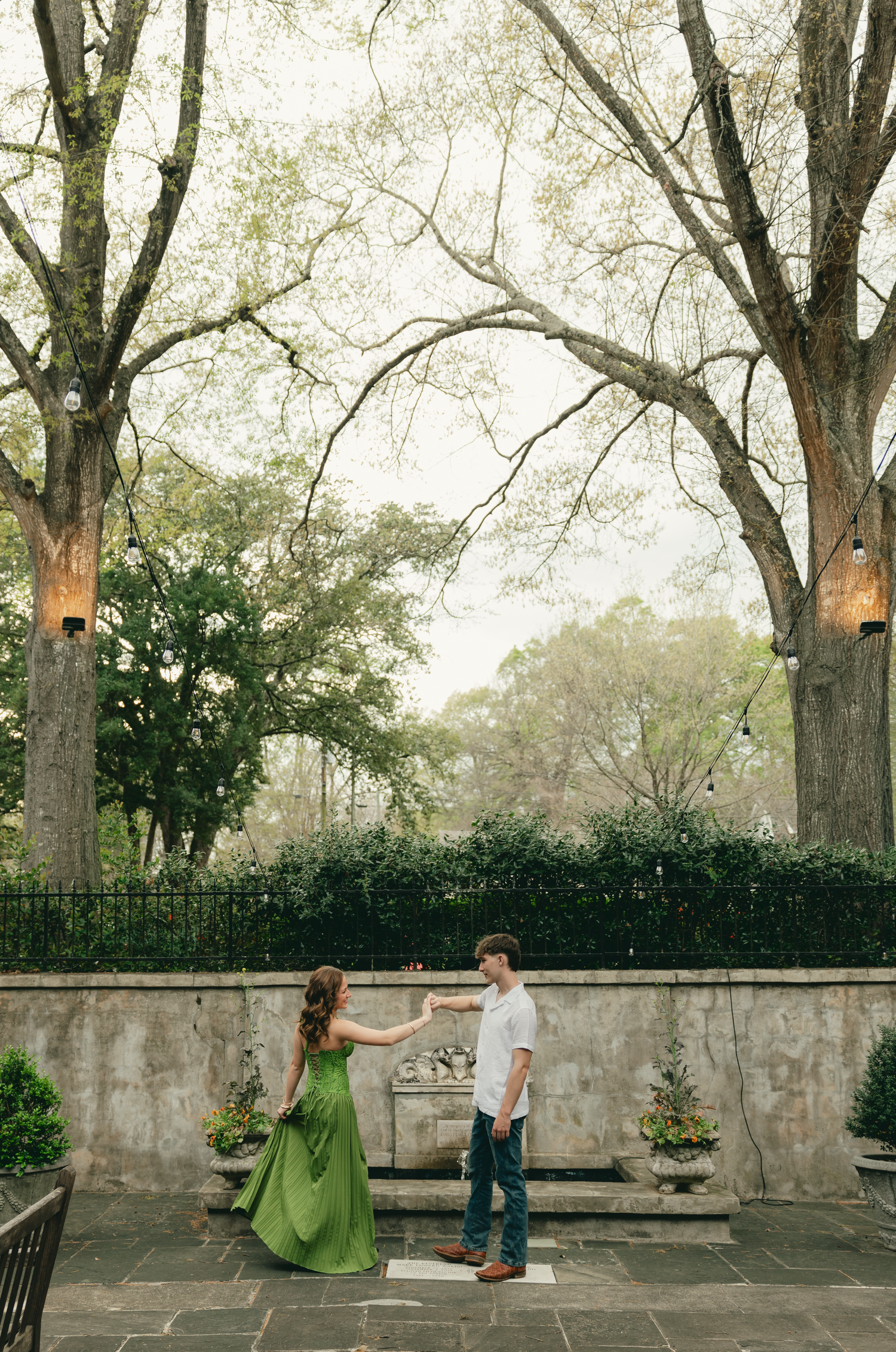 A couple dances together in a lush garden courtyard, the woman wearing a flowing green dress among tall trees.