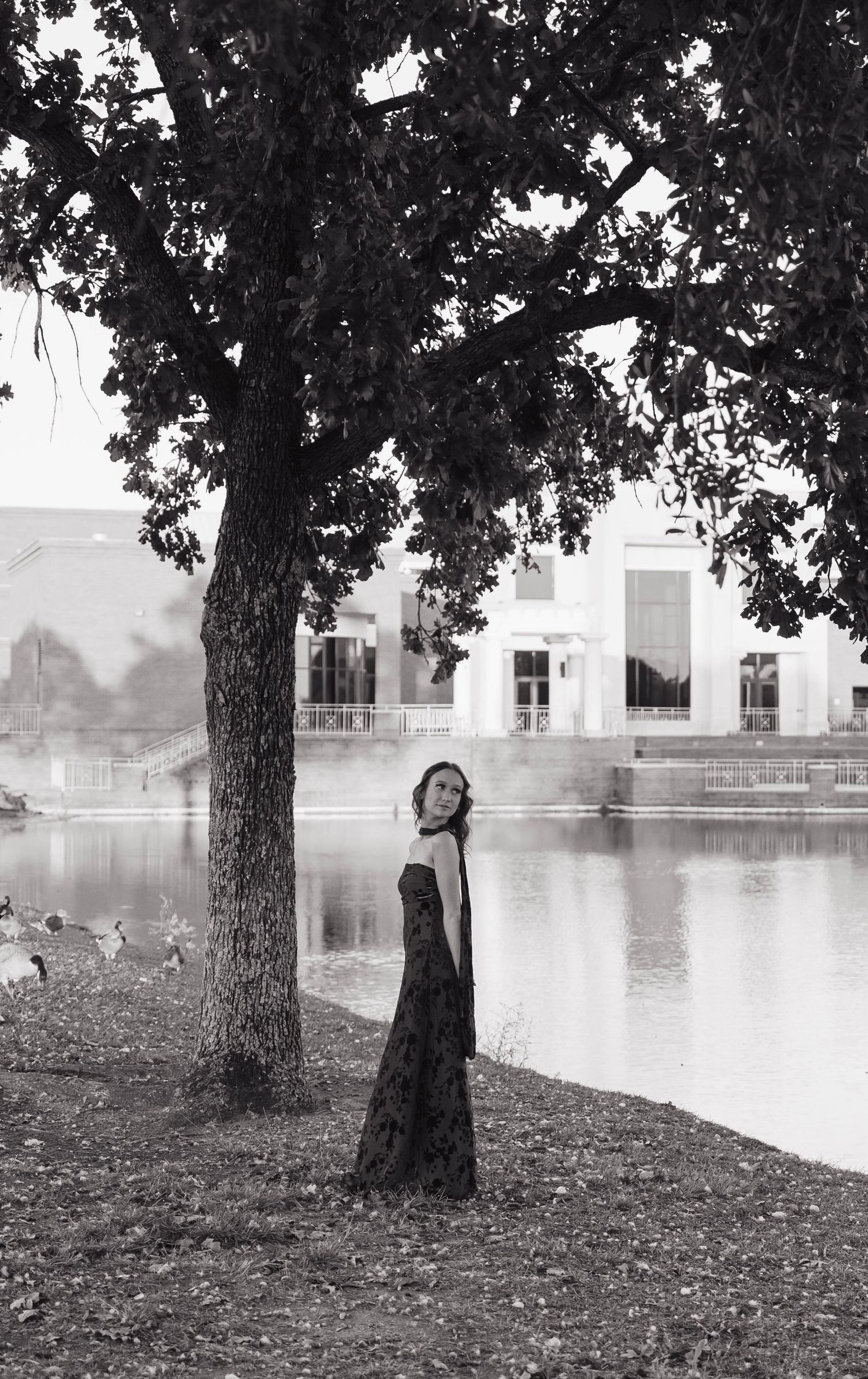 Black and white photo of a woman in a long dark dress standing by a tree near a lake with a building behind her.