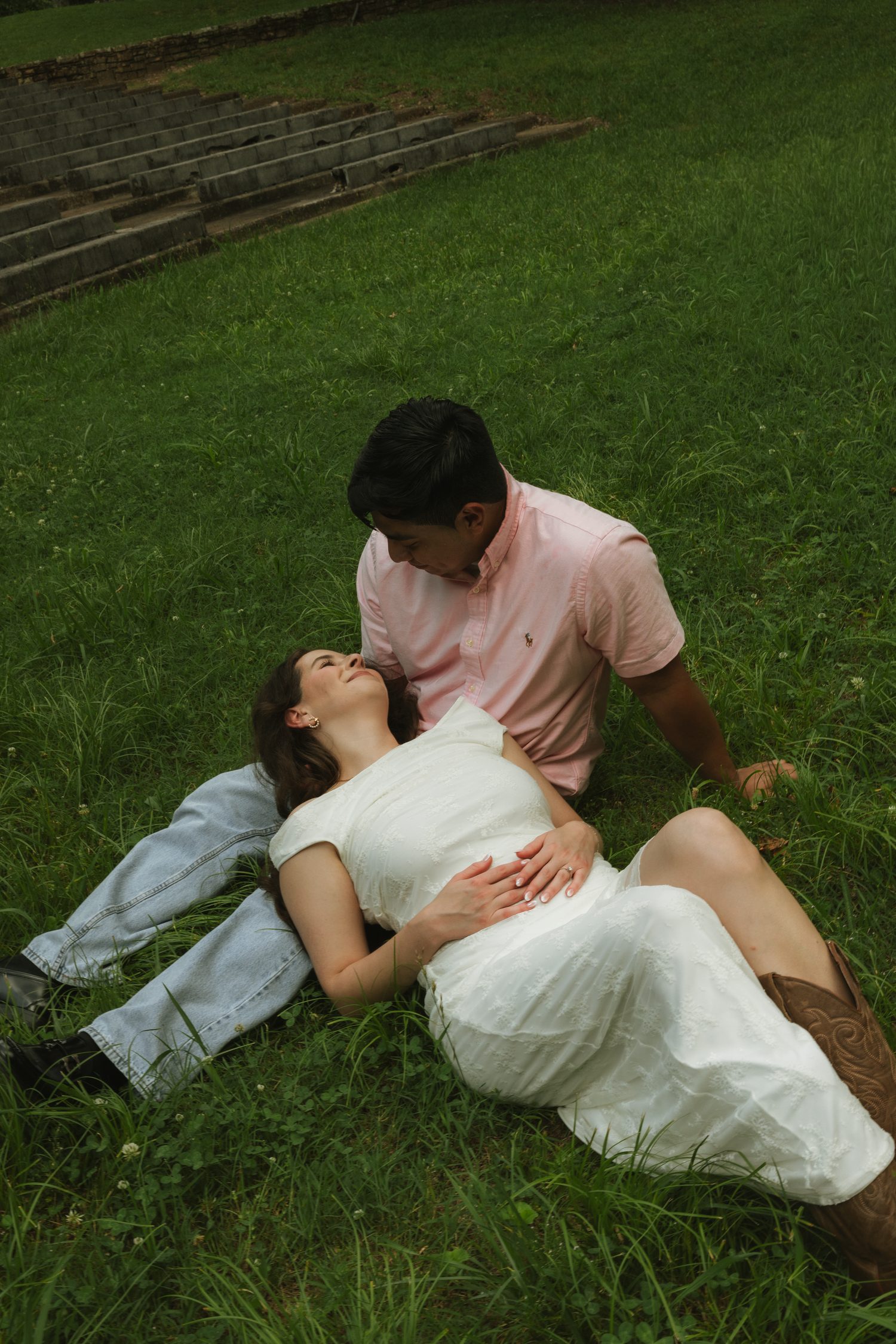 A pregnant woman in a white dress lies on green grass while her partner in a pink shirt tenderly touches her belly.