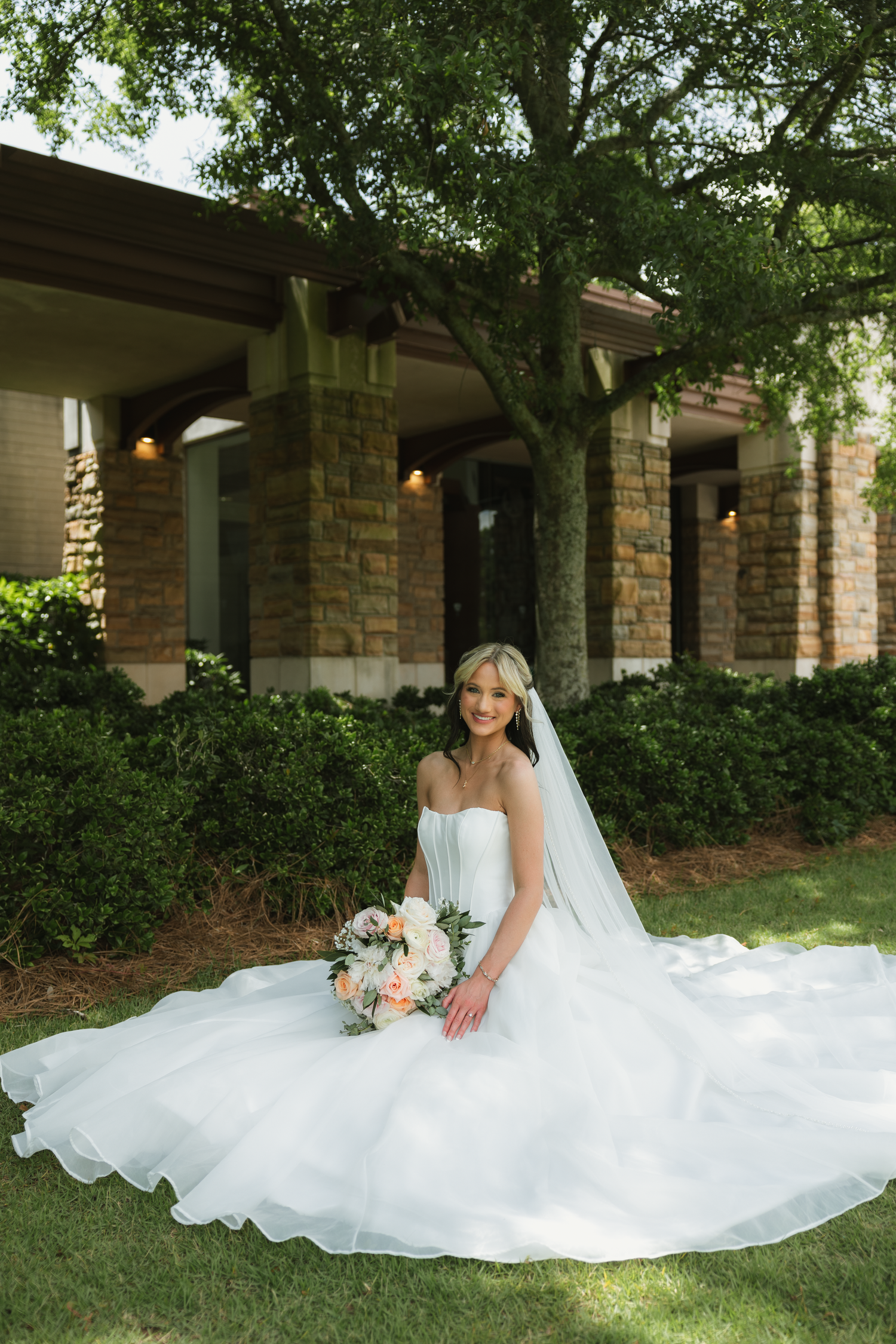 Bride in a white strapless ball gown with veil sits on grass holding a bouquet outside a stone building.