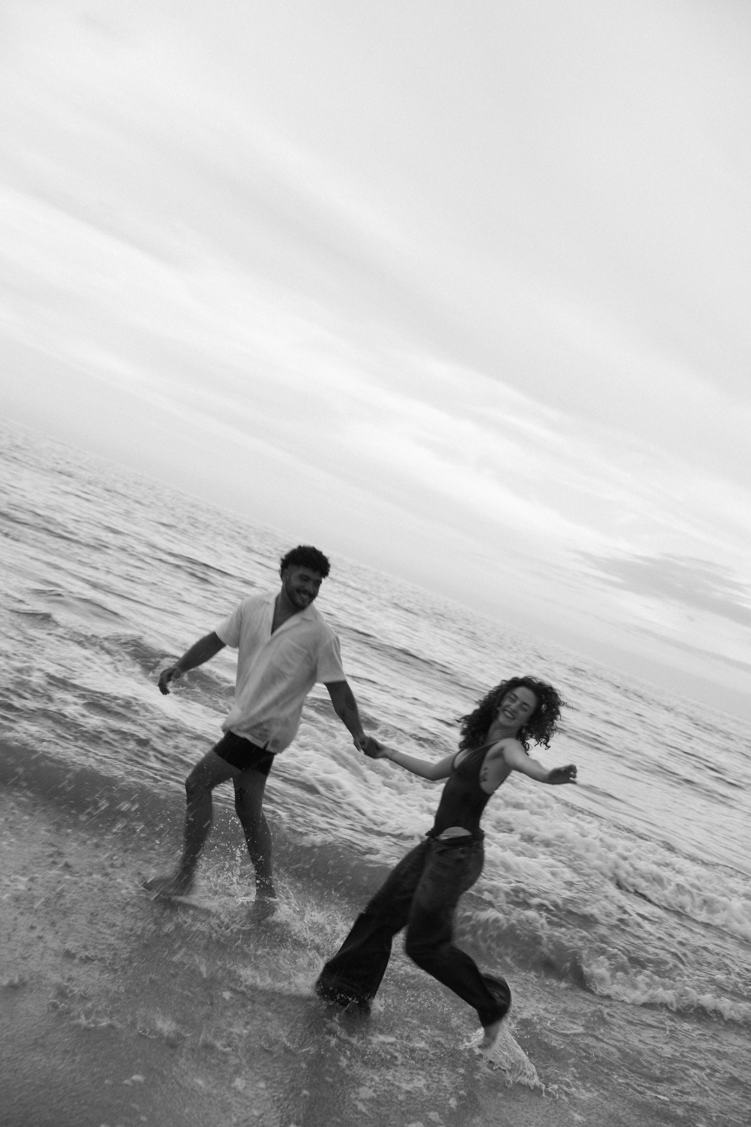 Black and white photo of a couple holding hands and running through ocean waves on a beach.