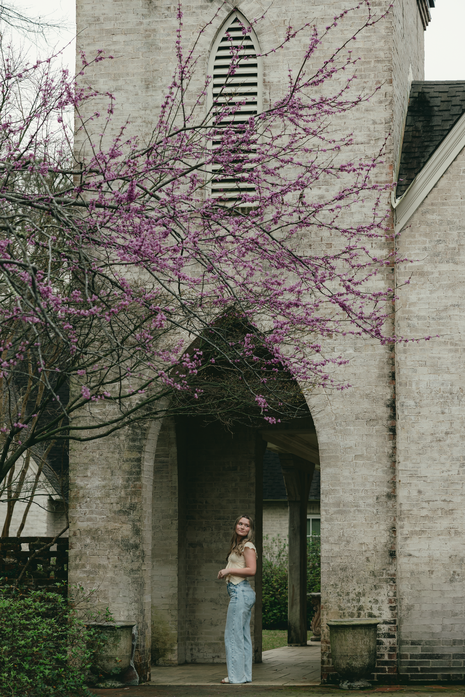 Woman in jeans stands under stone church arch with blooming pink redbud tree in spring.