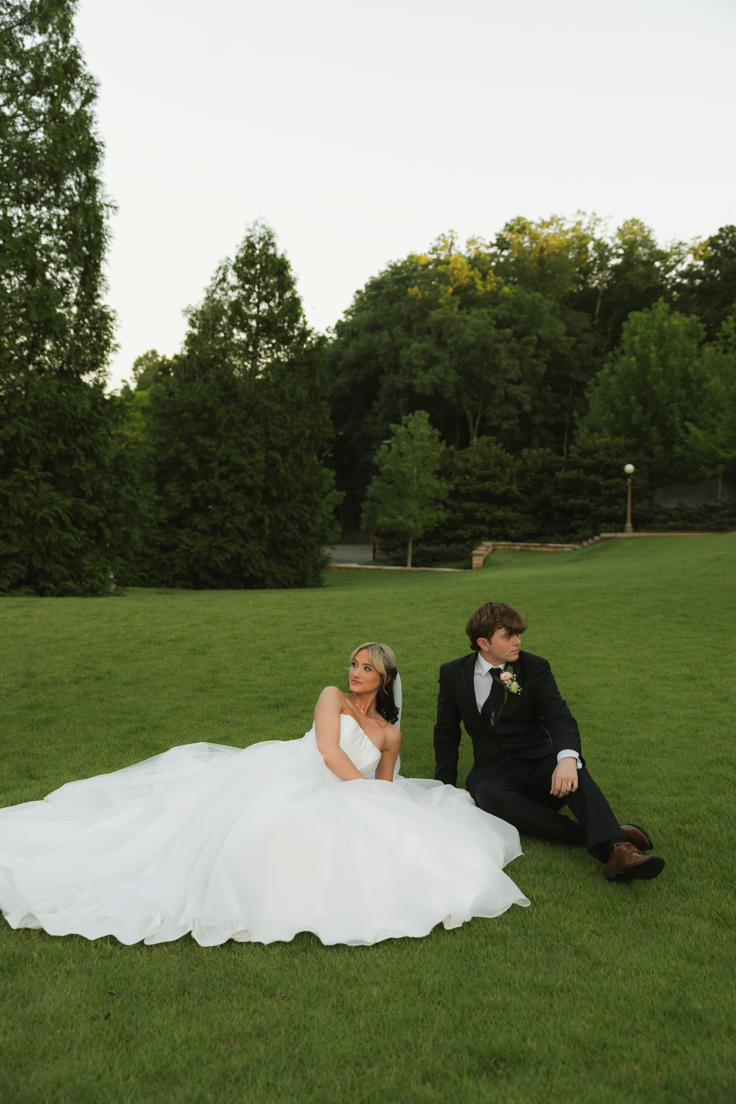 A bride in a white ballgown and groom in a black tuxedo sit together on lush green grass surrounded by trees.