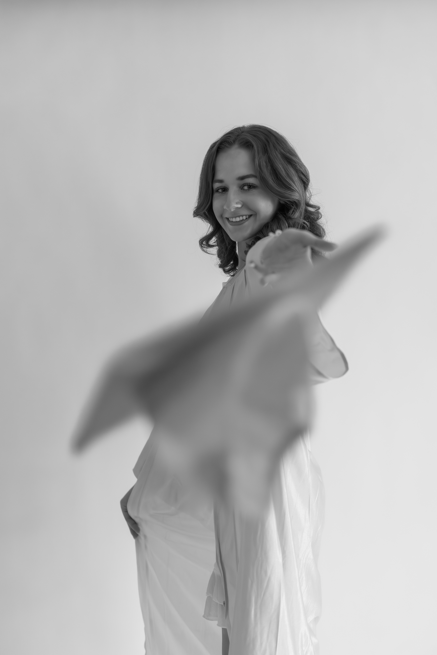 Black and white portrait of a smiling woman in white outfit throwing a graduation cap toward camera in a bright studio.