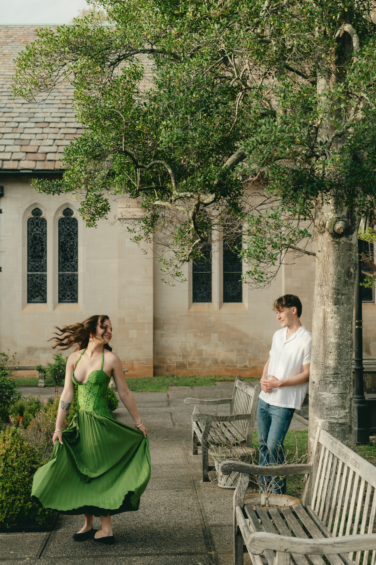 Couple in a garden courtyard, woman twirling in green dress, man in white shirt, stone church backdrop.