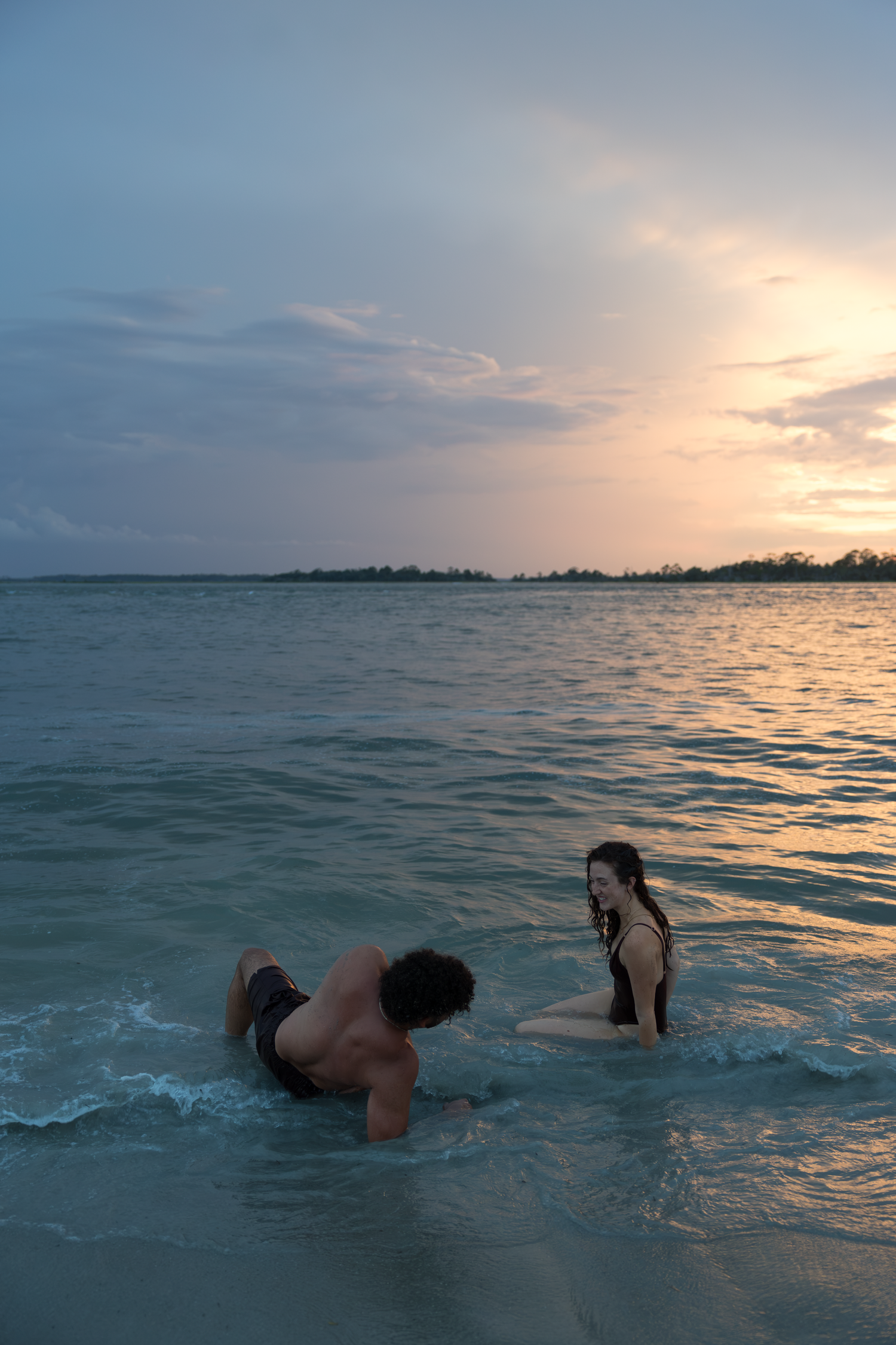 Couple playing in ocean waves at sunset with colorful sky and tropical shoreline in background.