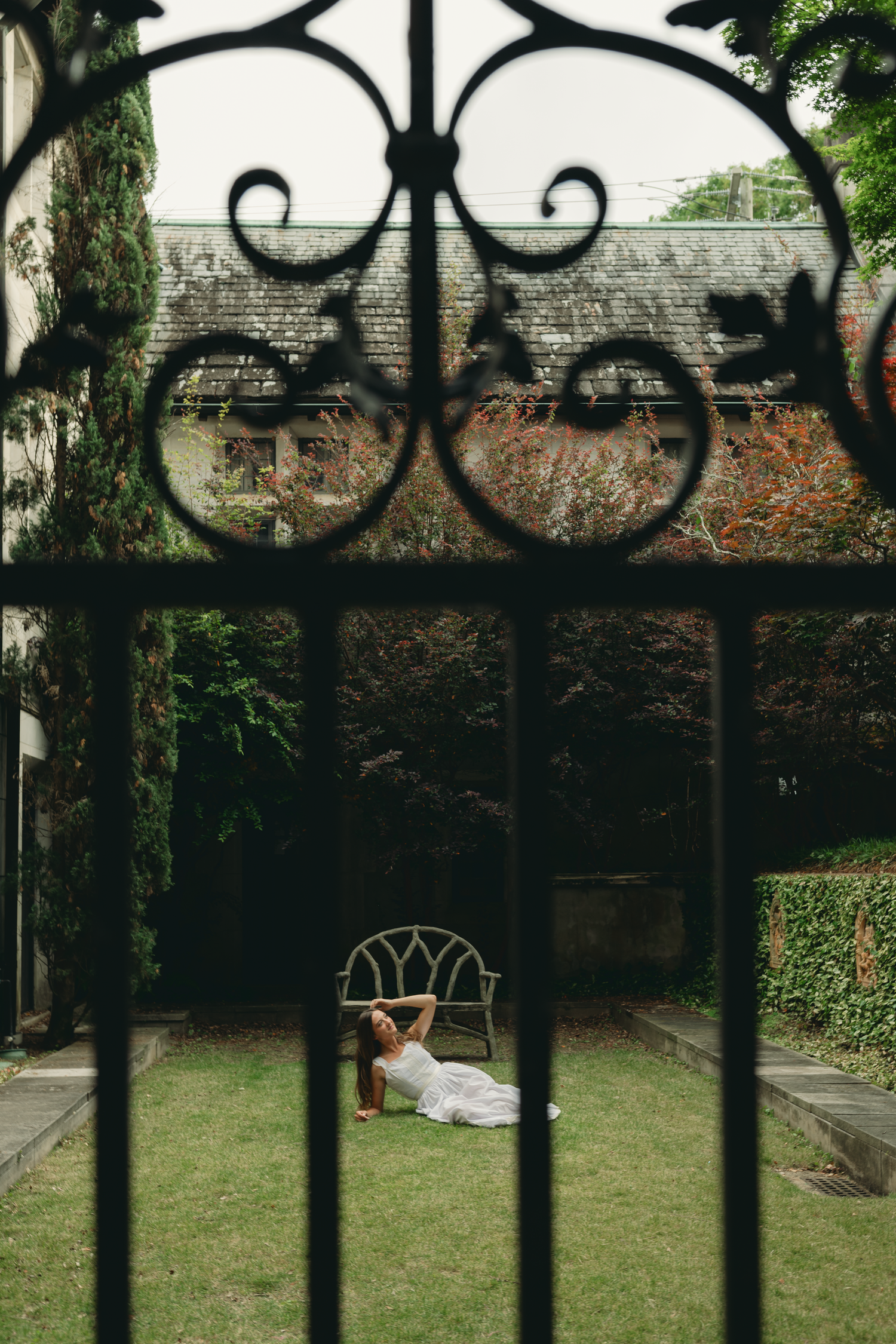 Woman in white dress lying on grass in a lush garden, viewed through ornate black iron gate.