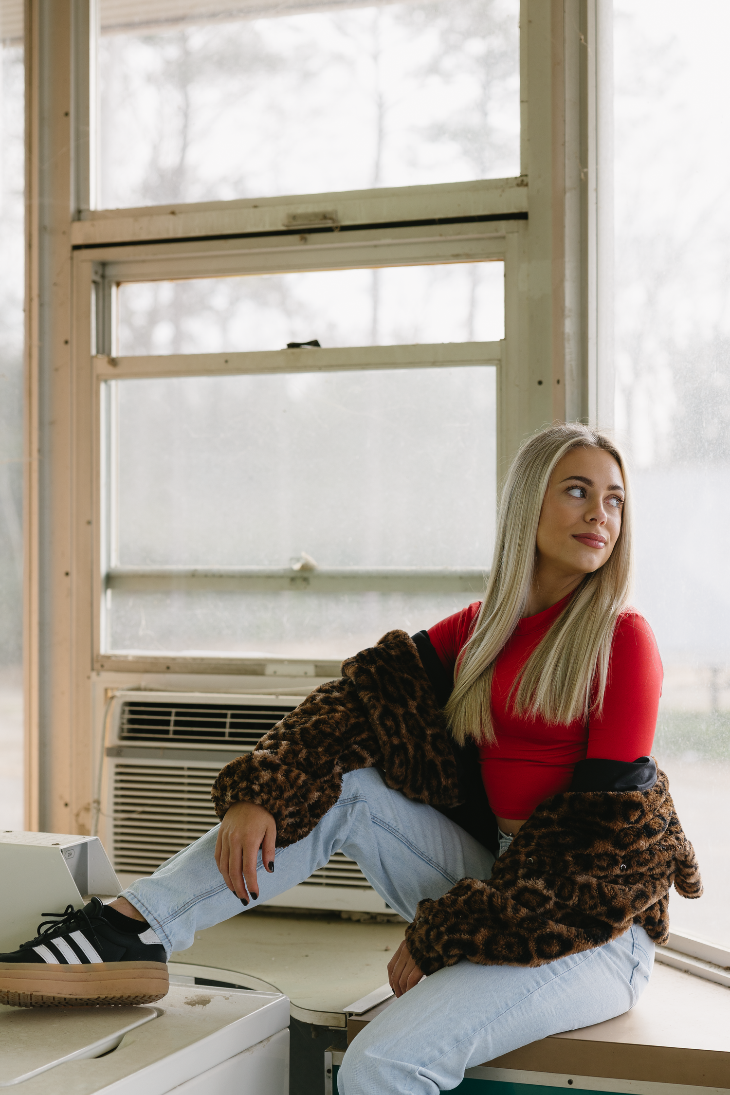 Blonde woman in red turtleneck, leopard coat, and jeans sitting on a counter near a window.