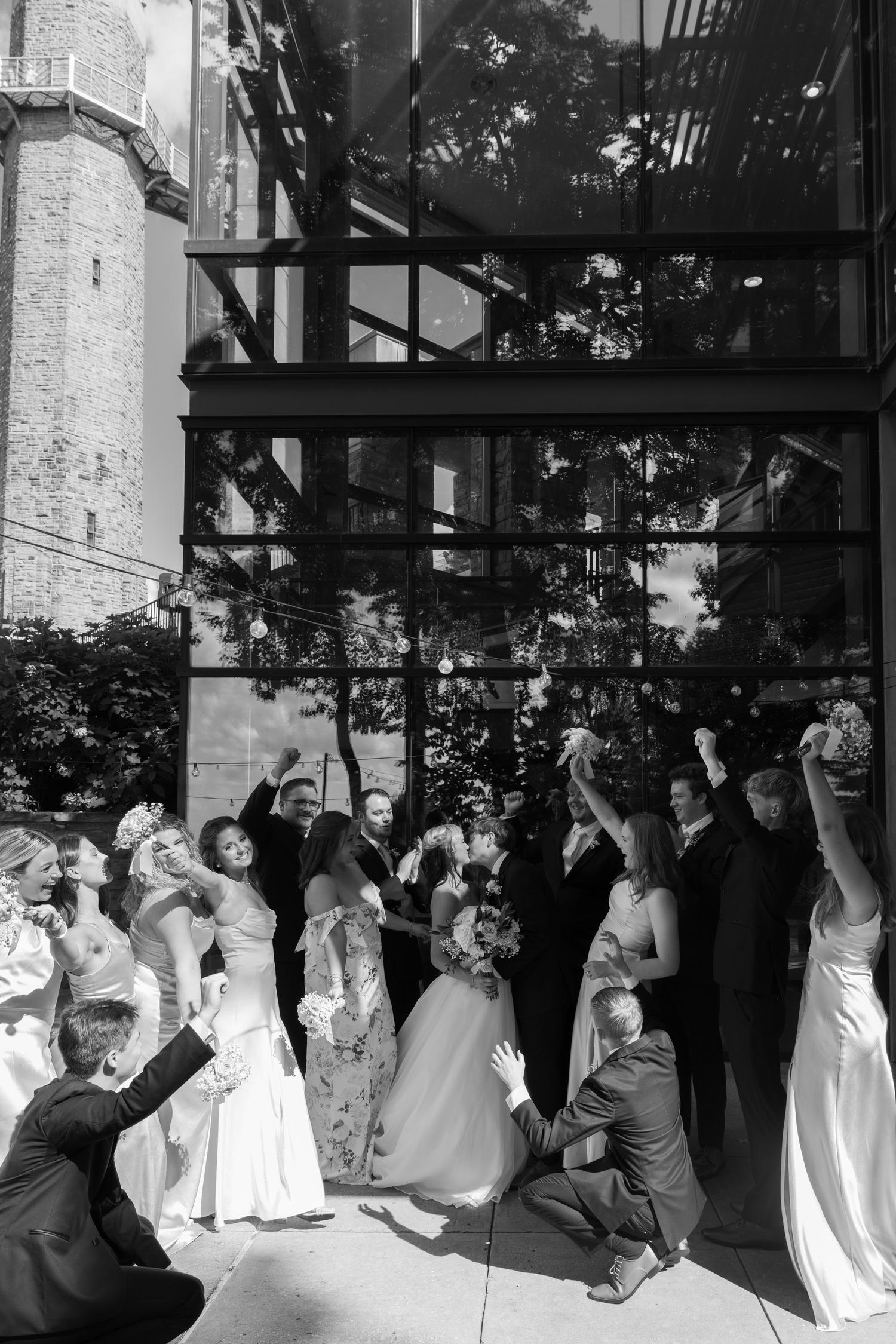 Black and white wedding party photo with bride, groom, and guests celebrating outdoors near a glass building.