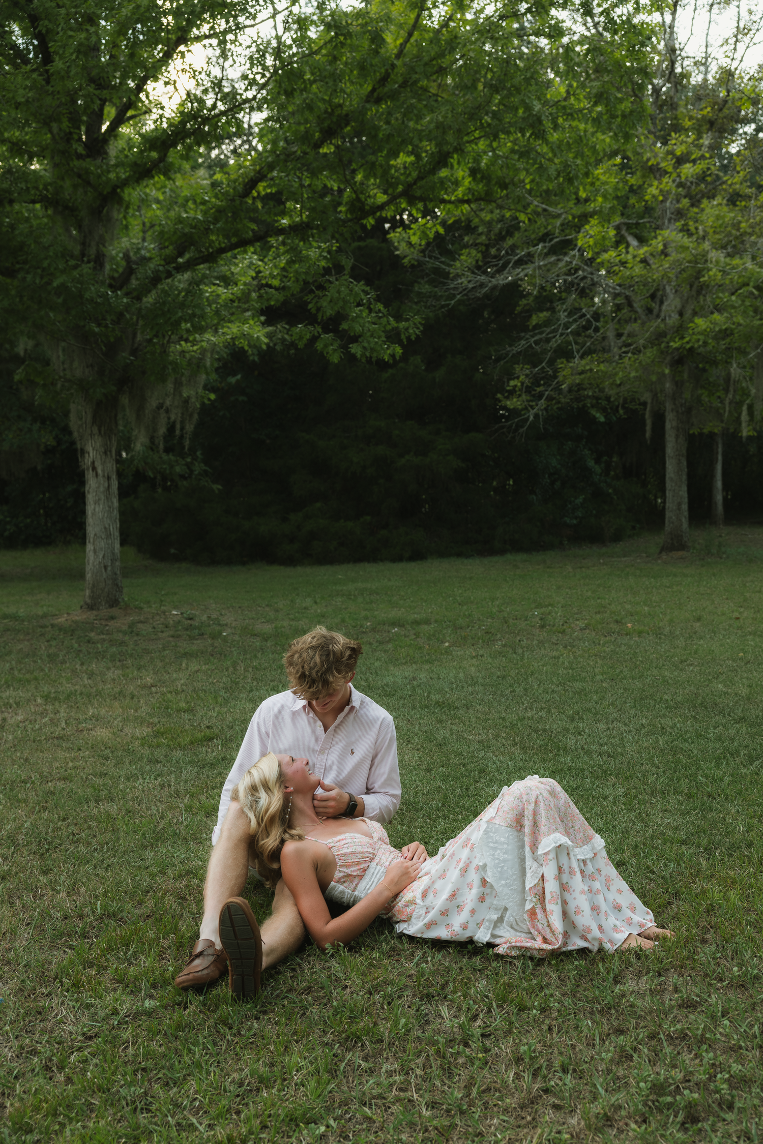 Couple relaxing on green grass in a park, woman lying in floral dress, man sitting beside her among lush trees.