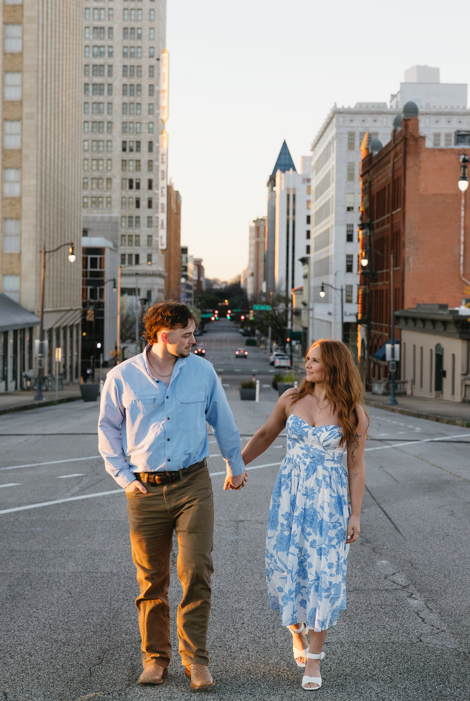 Couple holding hands walking in urban downtown street at sunset, woman in blue floral dress, man in light blue shirt.