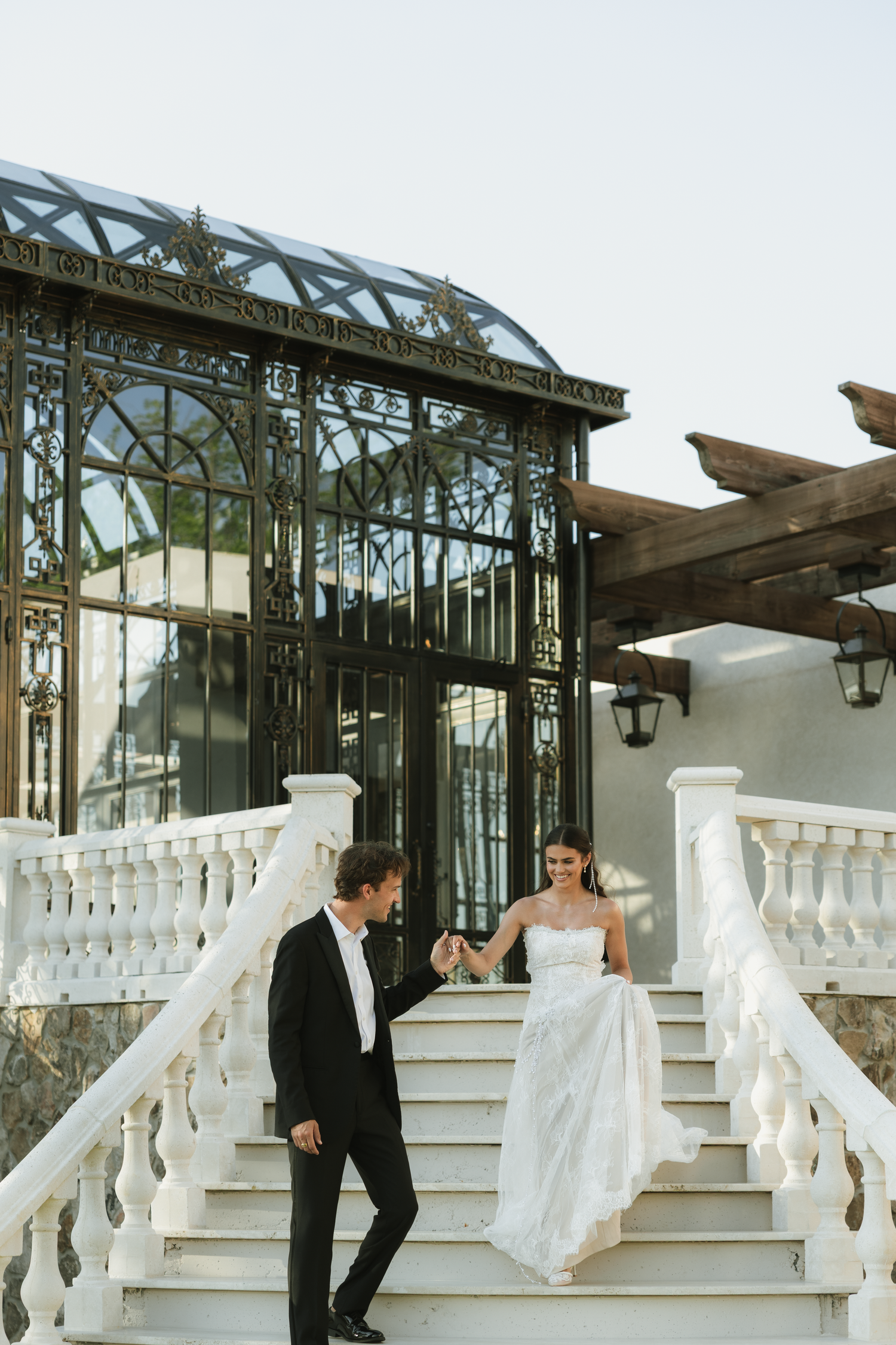 A bride and groom walk down elegant white balustrade stairs outside an ornate iron and glass greenhouse venue.