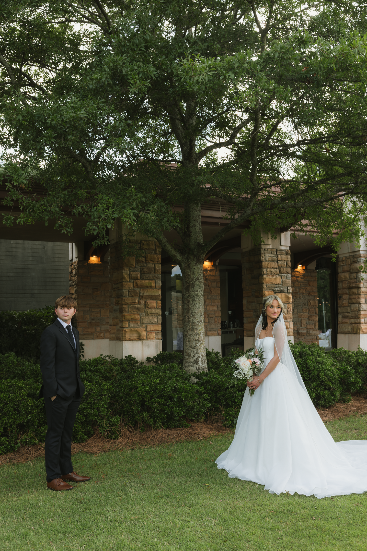 Bride in white gown holding bouquet stands apart from groom in black suit on green lawn before stone building.