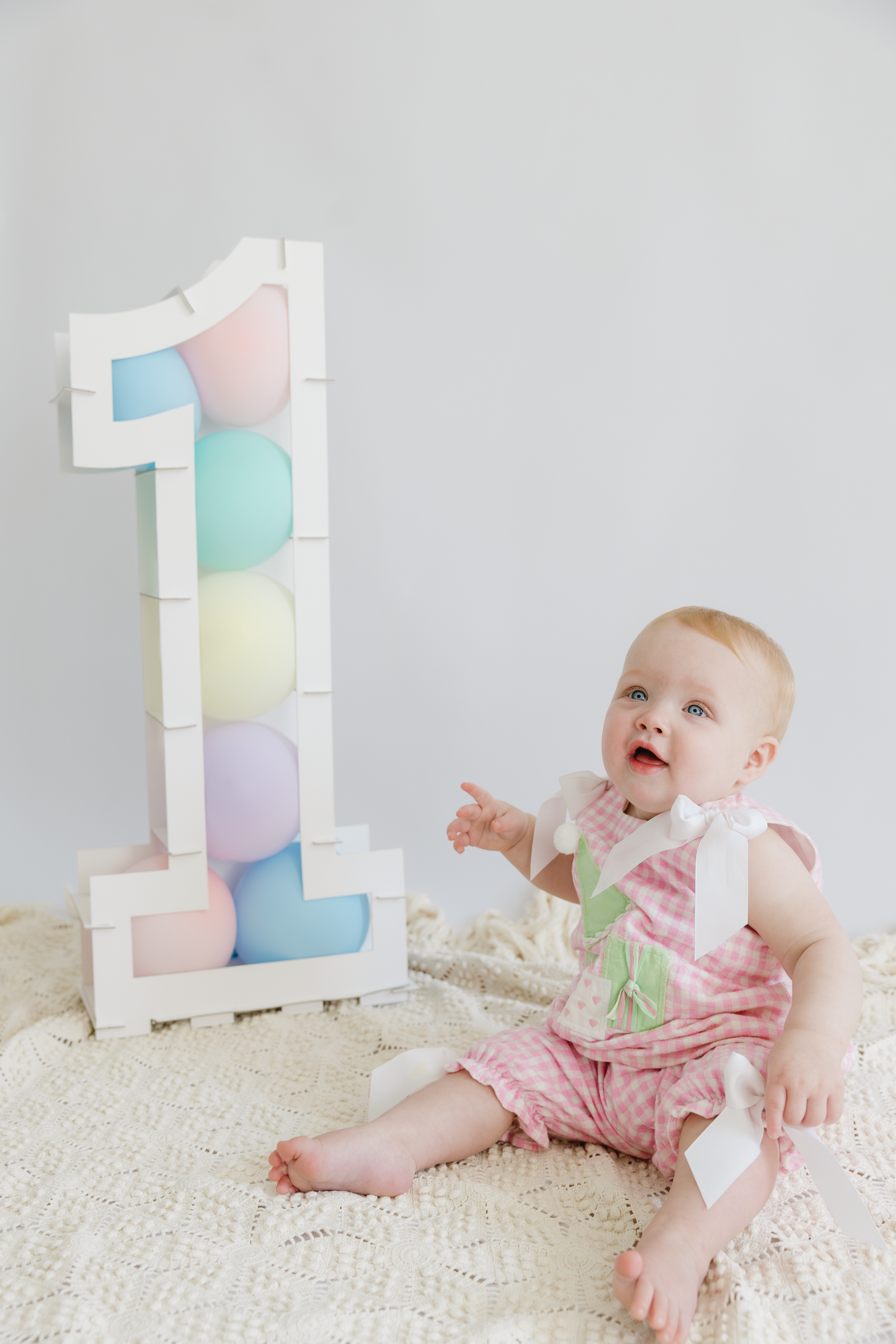 Baby girl in pink romper sits beside a large number 1 sign with pastel balloons celebrating her first birthday with a studio session.