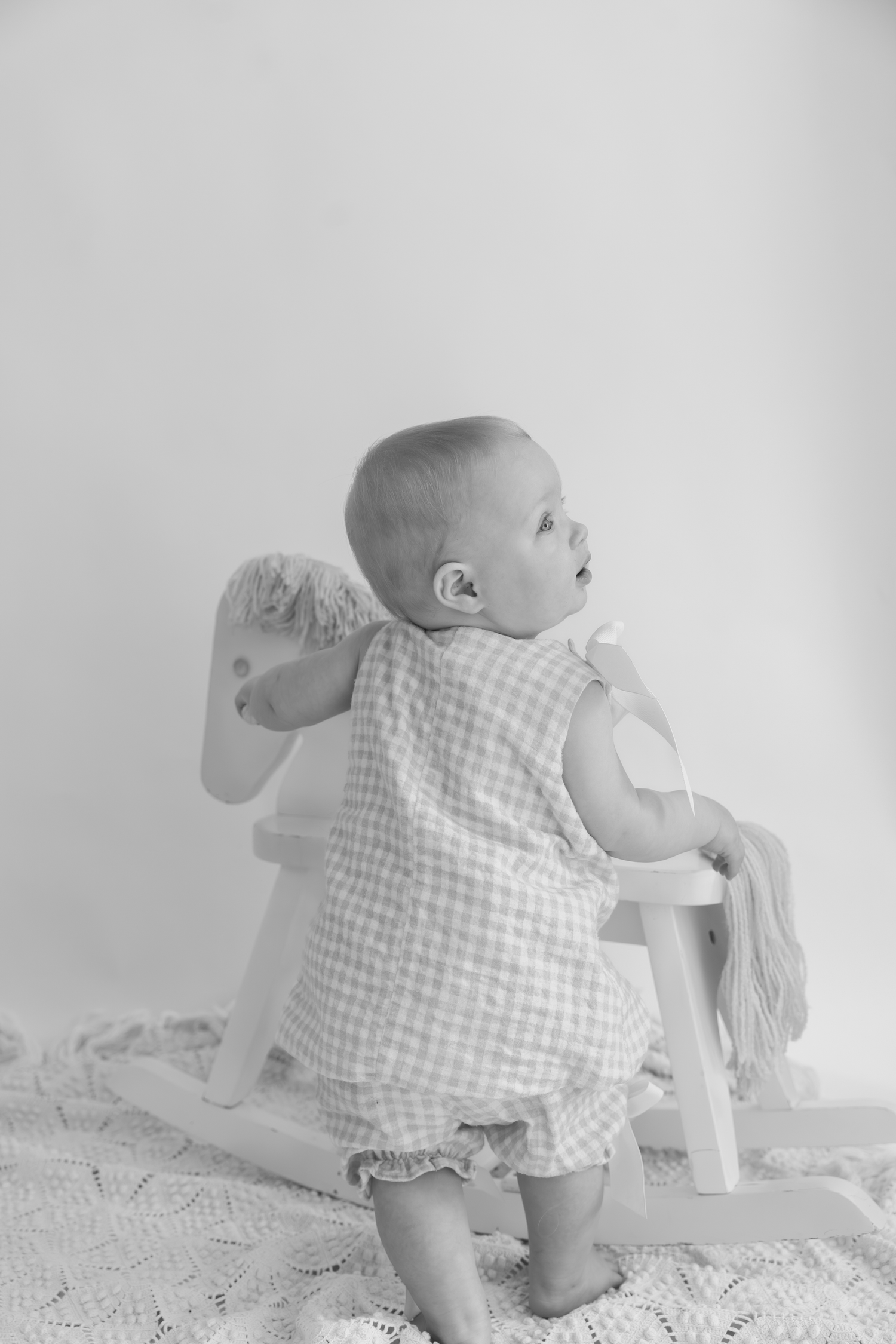 Black and white photo of a baby in gingham romper sitting on a small rocking horse, looking away.