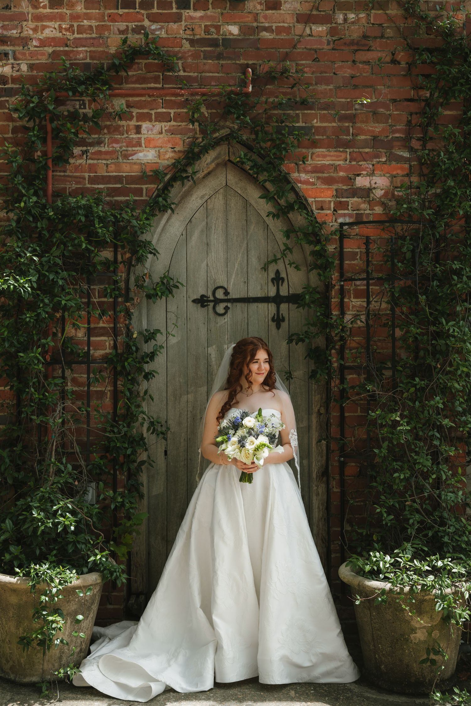 Bride in white ball gown holding floral bouquet stands before rustic wooden arched door framed by ivy on brick wall.