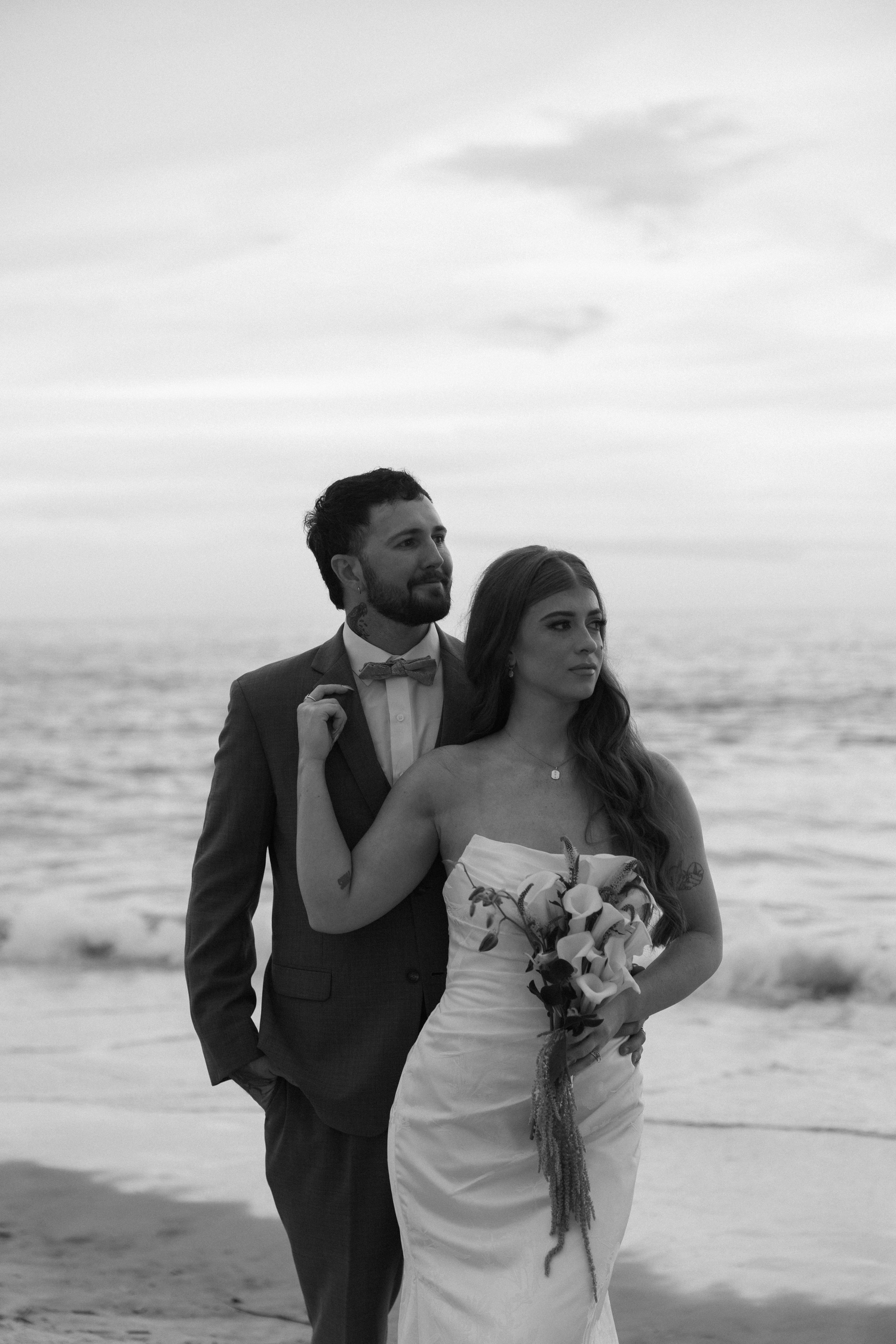 Black and white beach wedding photo of bride holding bouquet and groom in suit embracing by the ocean at dusk.