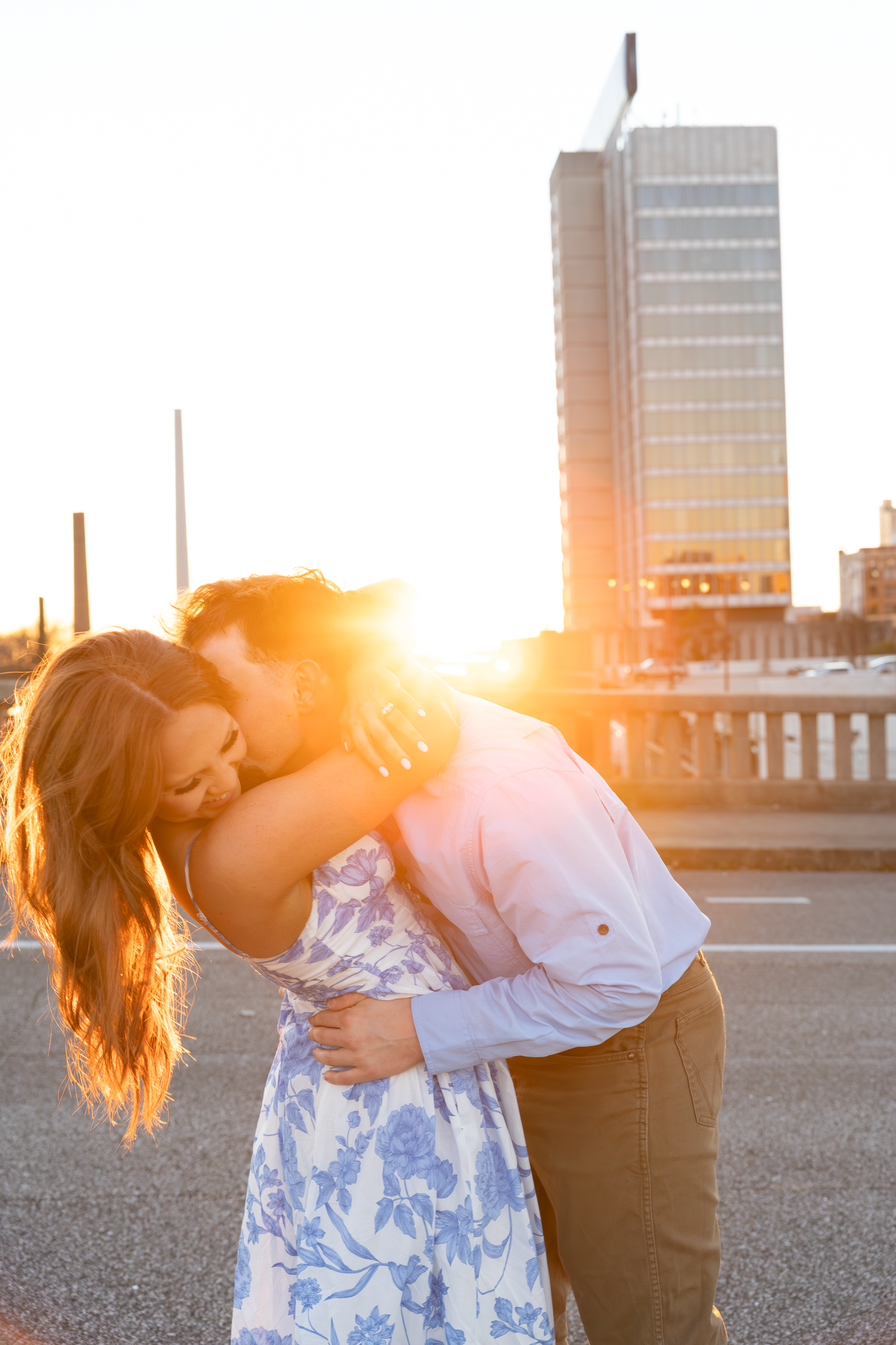 Couple embracing romantically at sunset with golden light and city skyline in the background.