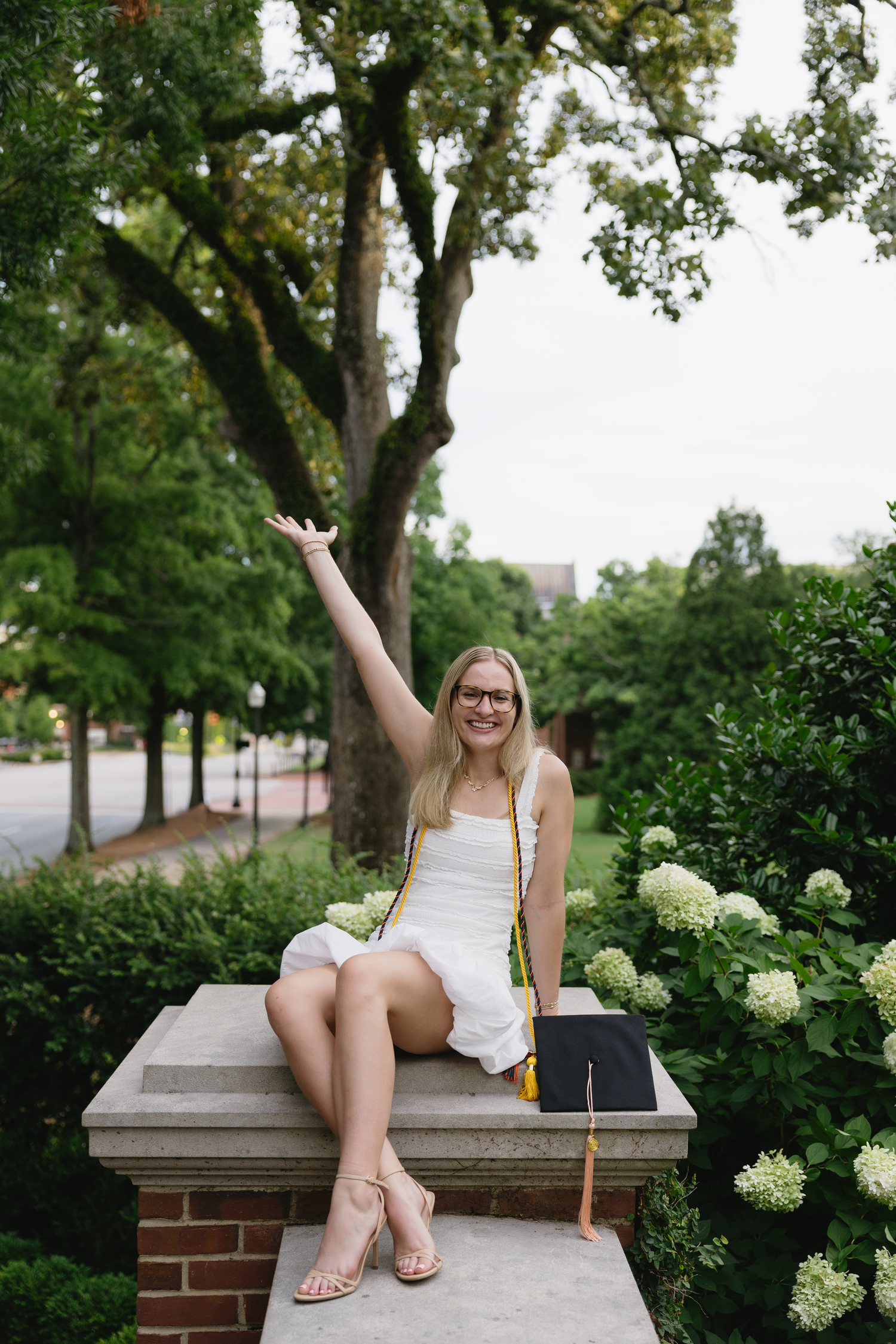 A smiling blonde woman in a white dress celebrates graduation, sitting outdoors with her cap and colorful tassel.