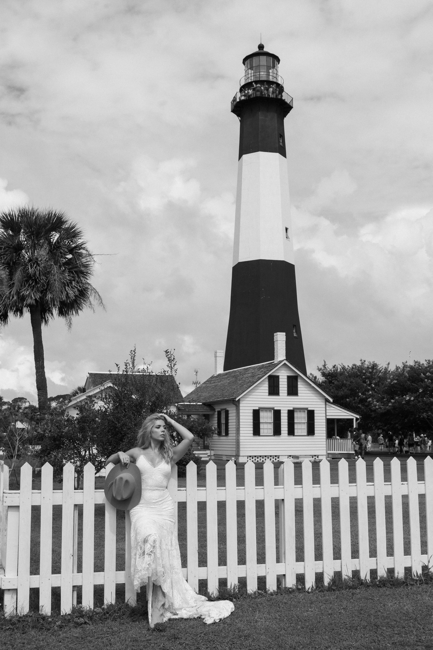 Black and white photo of a bride in lace gown posing by white picket fence with striped lighthouse and palm trees.