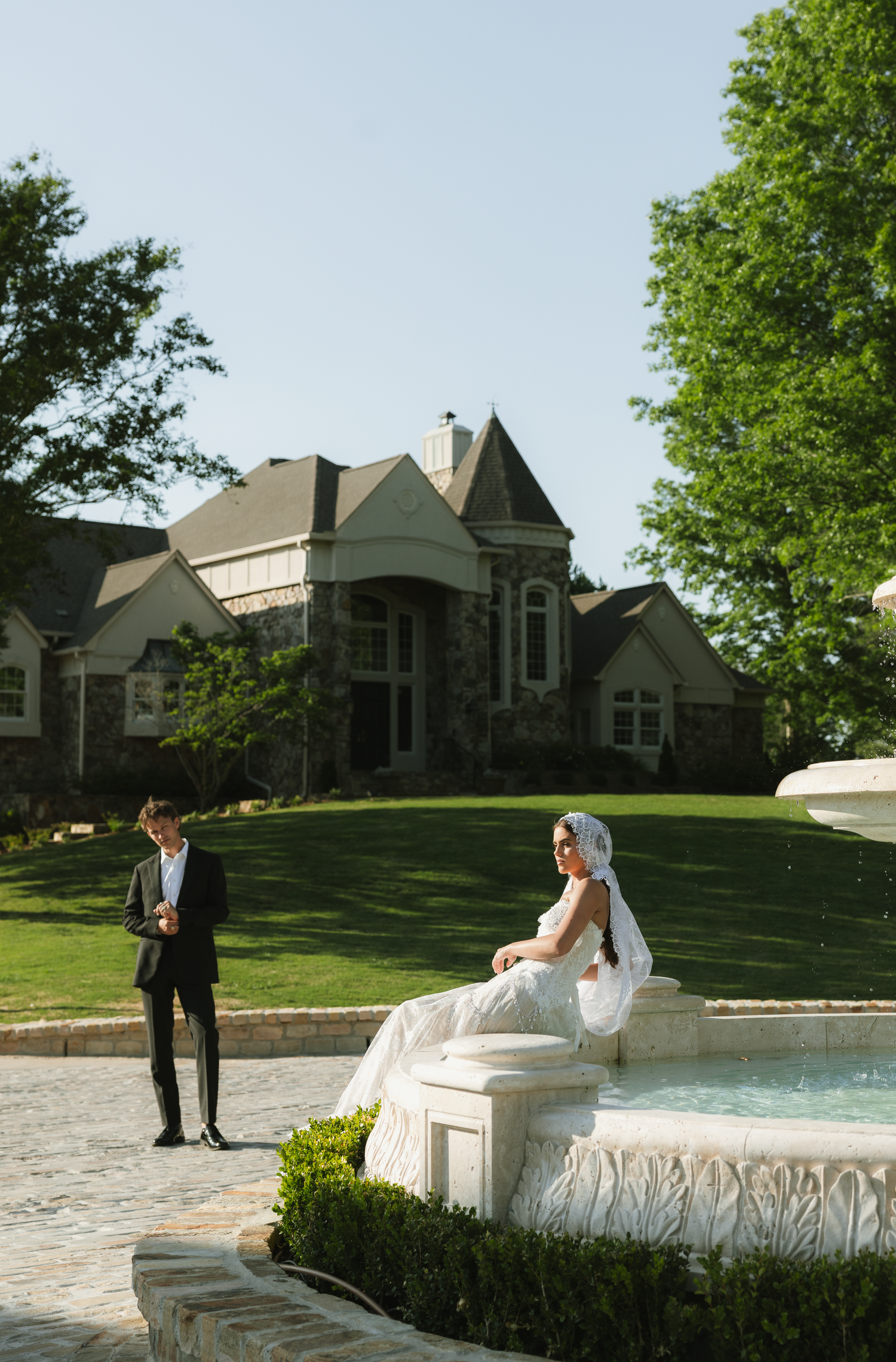 A bride in a white gown sits on a fountain while a groom in a suit stands nearby at an elegant estate.