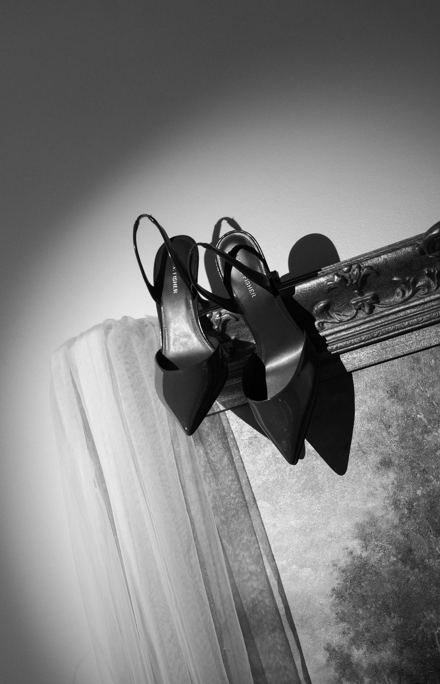 Black slingback heels resting on an ornate mirror frame beside a white veil in elegant black and white photography.