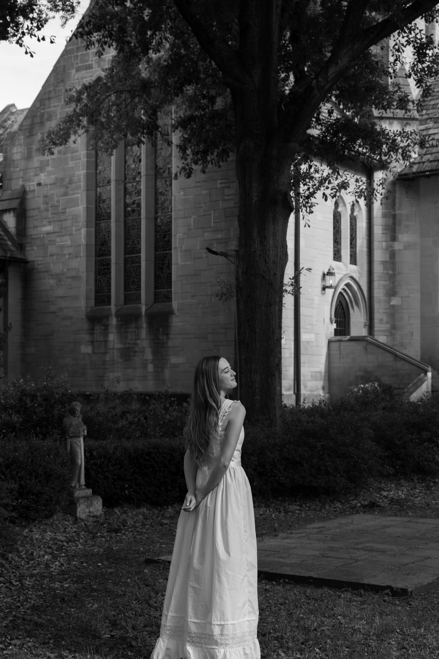 Black and white photo of a woman in a white dress standing by a large tree near a historic stone church.
