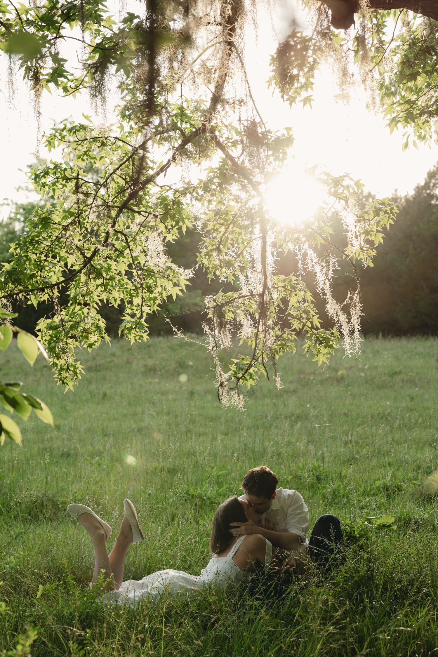 Romantic couple lying together in lush green meadow under sunlit trees, sharing an intimate moment outdoors.