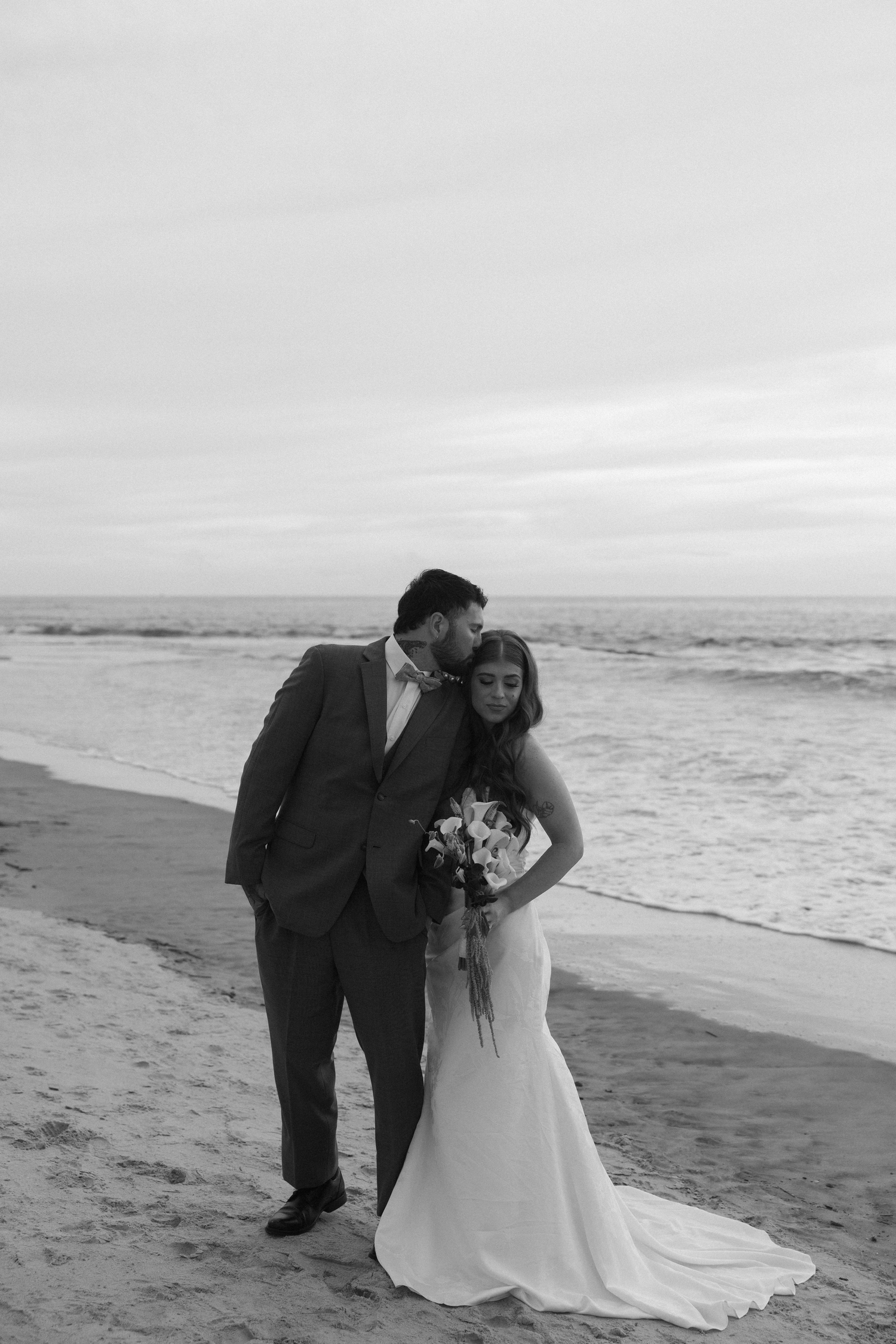 Black and white beach wedding photo of groom kissing bride on cheek while she holds a bouquet by the ocean.